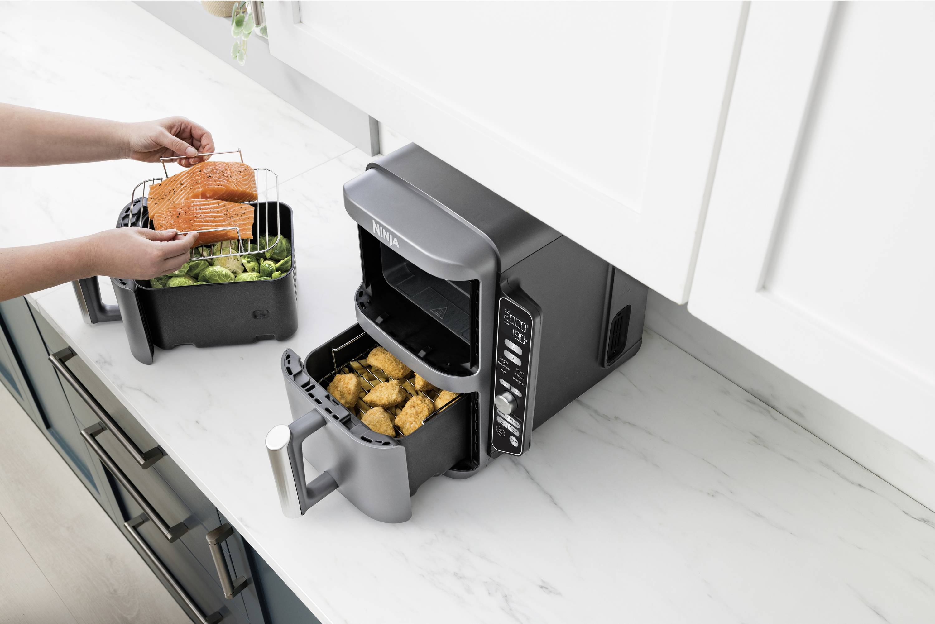 A person is placing salmon fillets on a metal grid in an air fryer basket, with vegetables cooking alongside. Next to it is another air fryer with chicken nuggets.