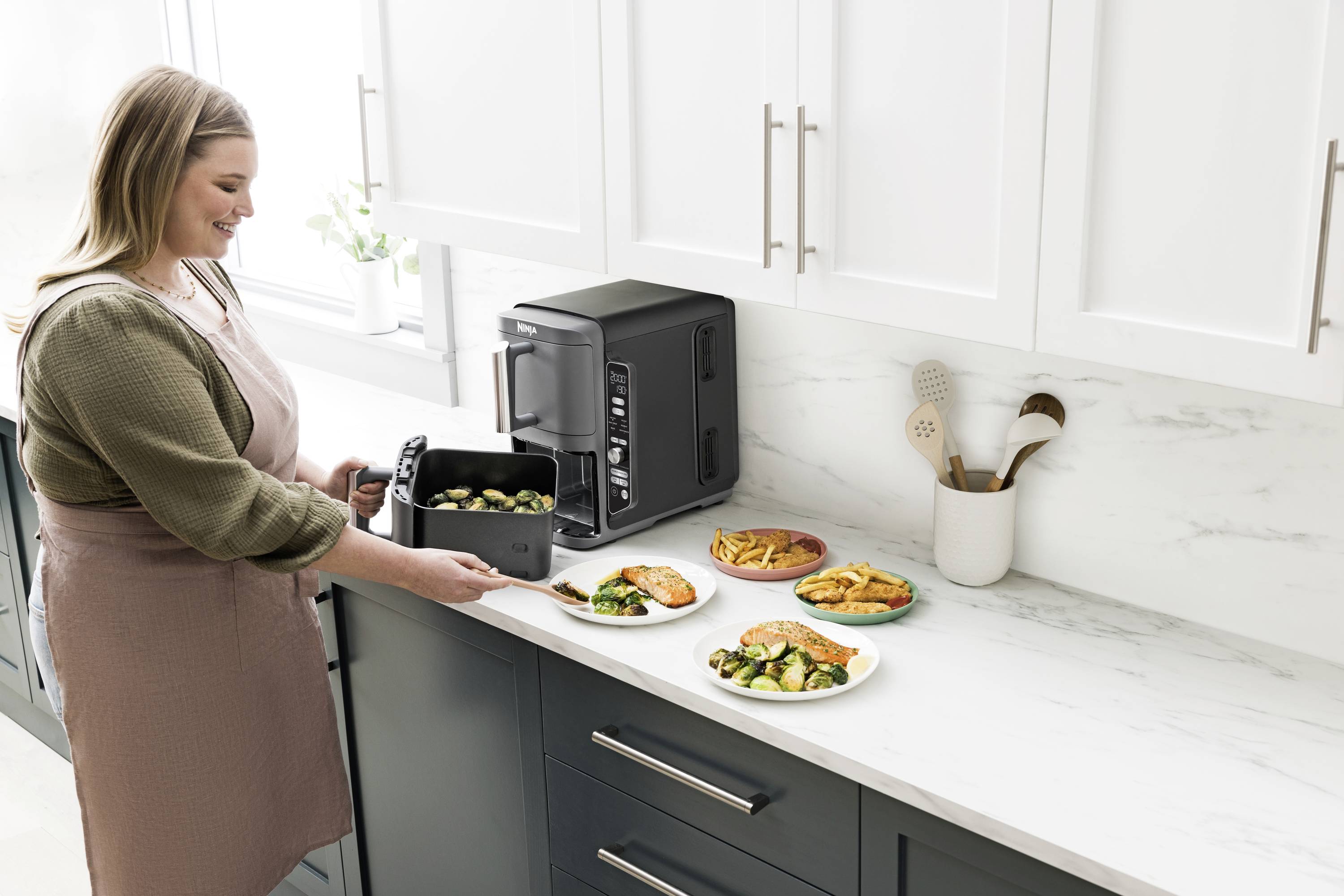 A woman in a kitchen is taking food out of an air fryer and placing it onto plates. Several dishes are standing on the worktop.