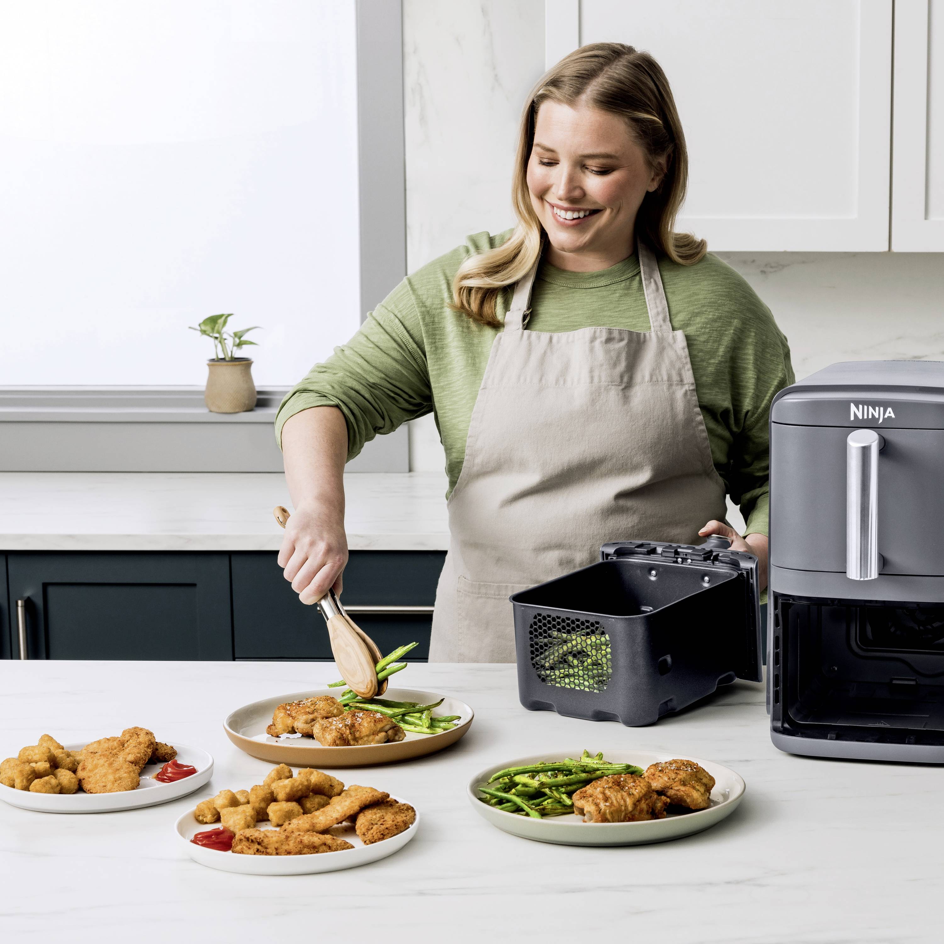 A woman serves baked food from an air fryer onto plates with green beans, in a modern kitchen.