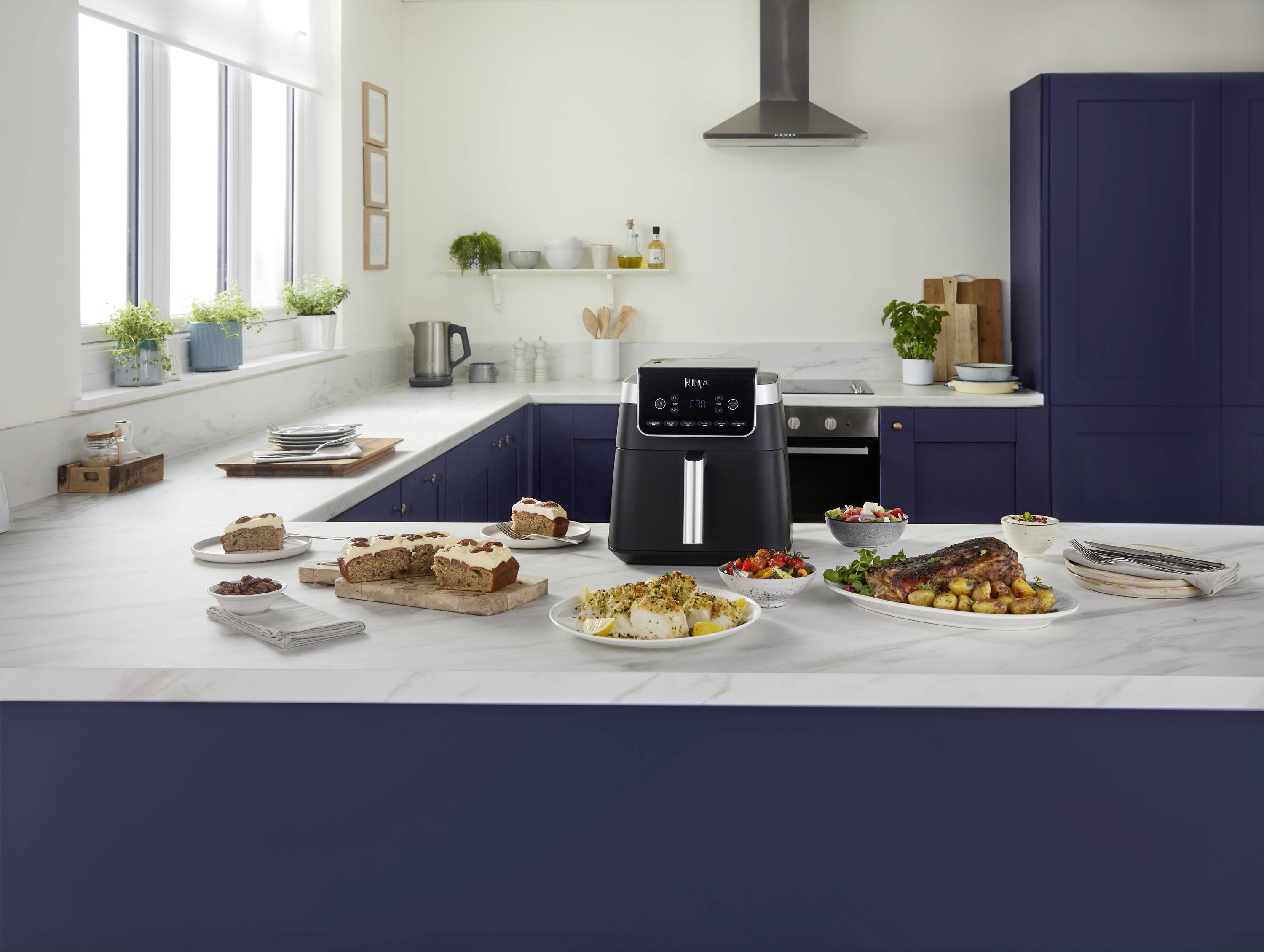 A modern kitchen with blue cabinetry. In the foreground, an air fryer sits on the worktop, surrounded by various dishes.