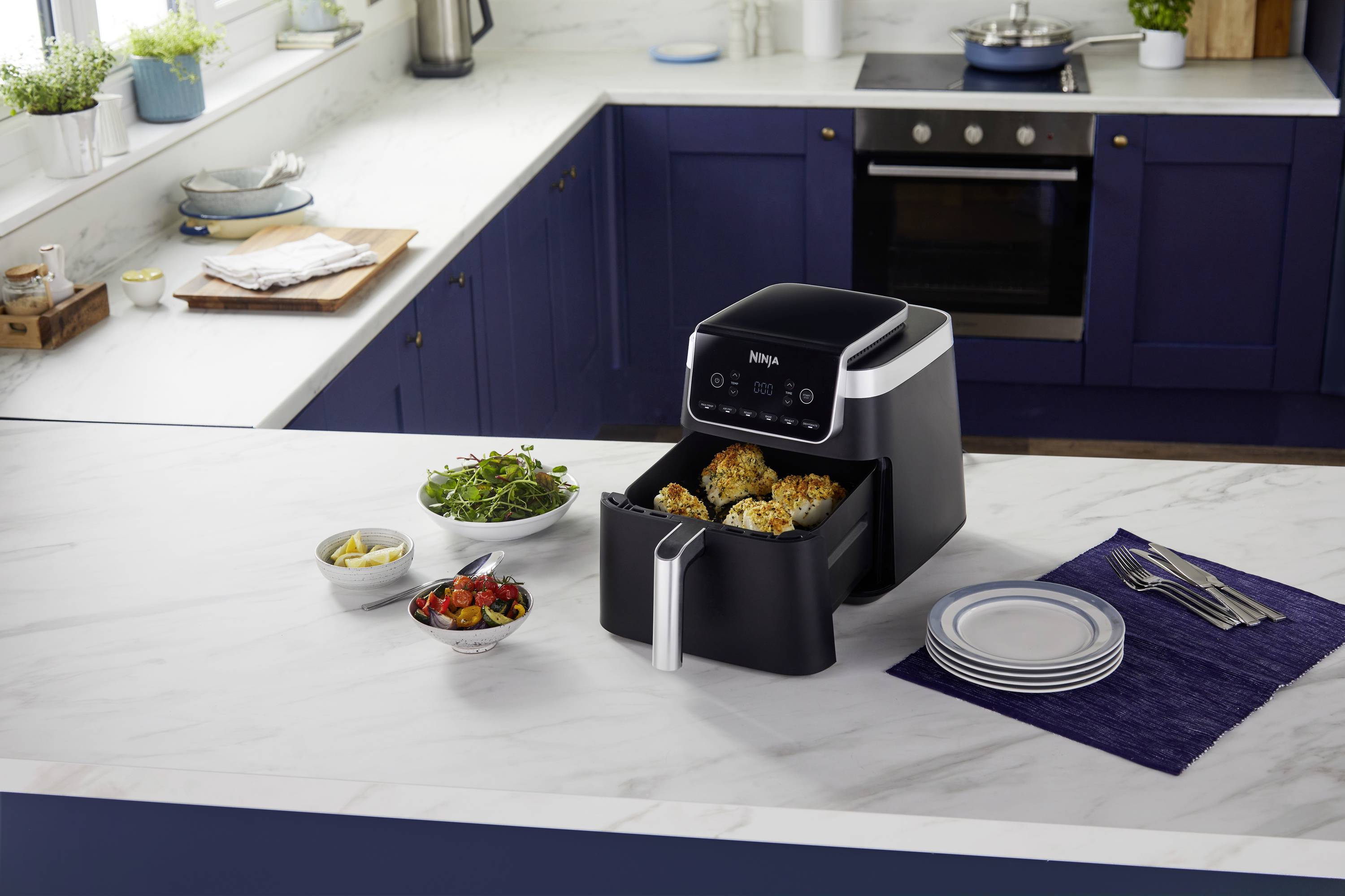 An air fryer sits on a worktop in a modern kitchen. The drawer is open and filled with vegetables.