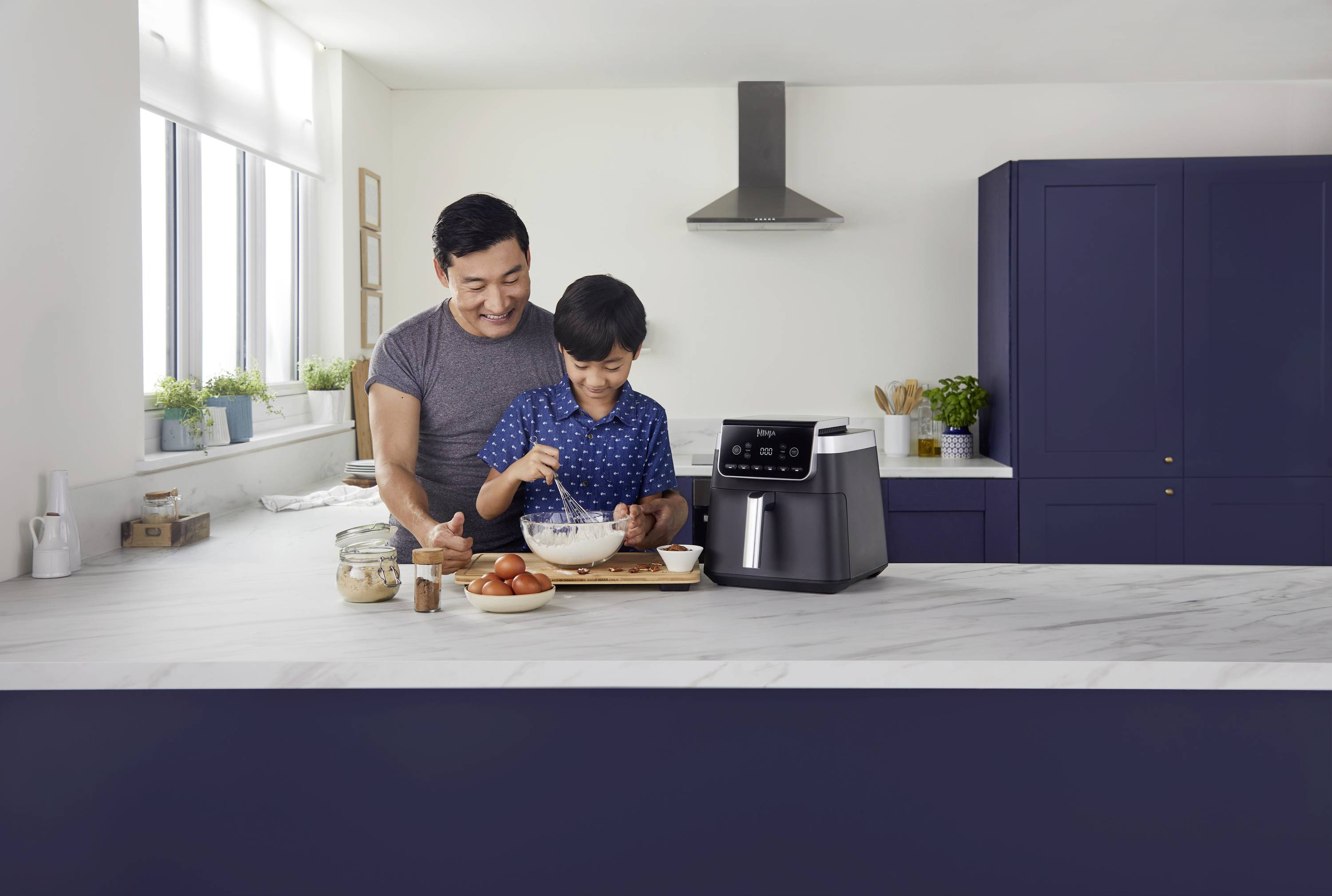 A man and a child are baking together in a modern kitchen. An air fryer sits on the worktop.