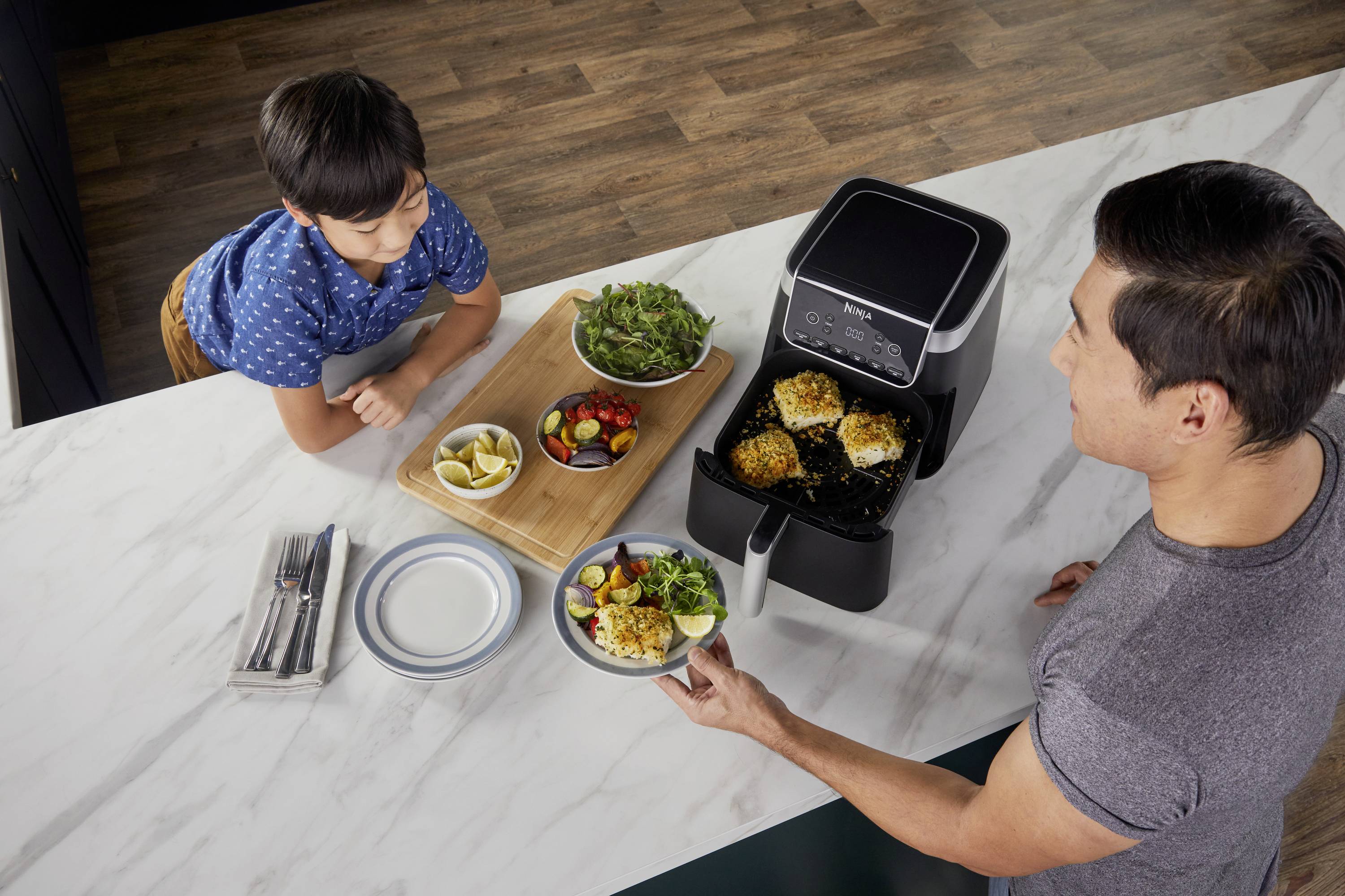 A man serves a dish from an air fryer to a boy at the table. Salad, lemons, and an empty plate set are on the table.