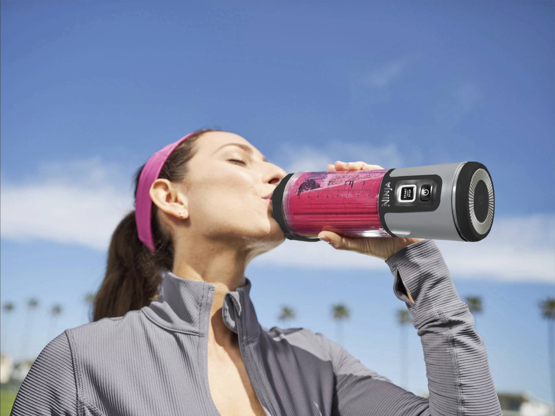 A woman is drinking from a portable blender containing a red beverage, with a blue sky in the background. She is wearing a headband and sportswear.