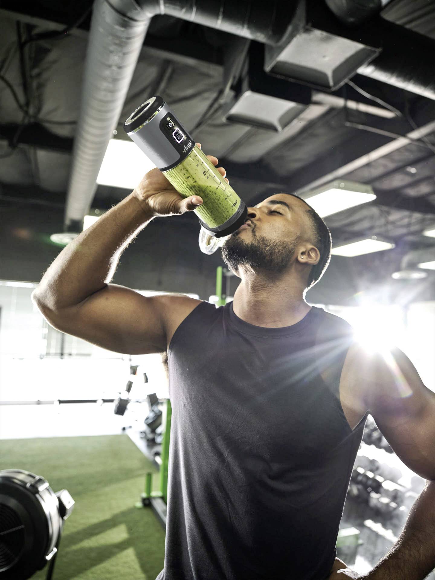 A man in a gym is drinking from a sports bottle. Fitness equipment can be seen in the background. Sunlight is streaming through the windows.