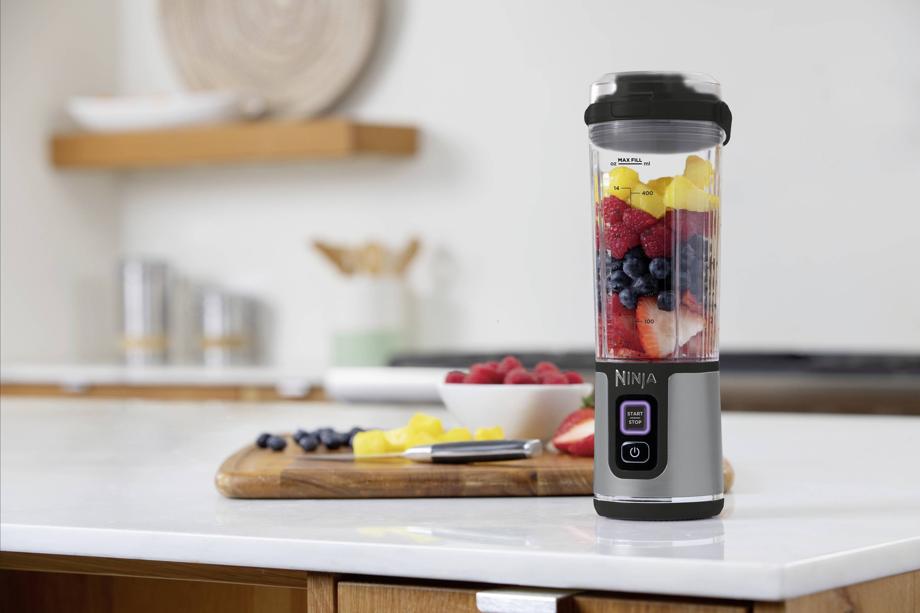 A blender sits on a kitchen worktop, filled with various fruits including strawberries, blueberries, and pineapple chunks. Kitchen utensils are visible in the background.