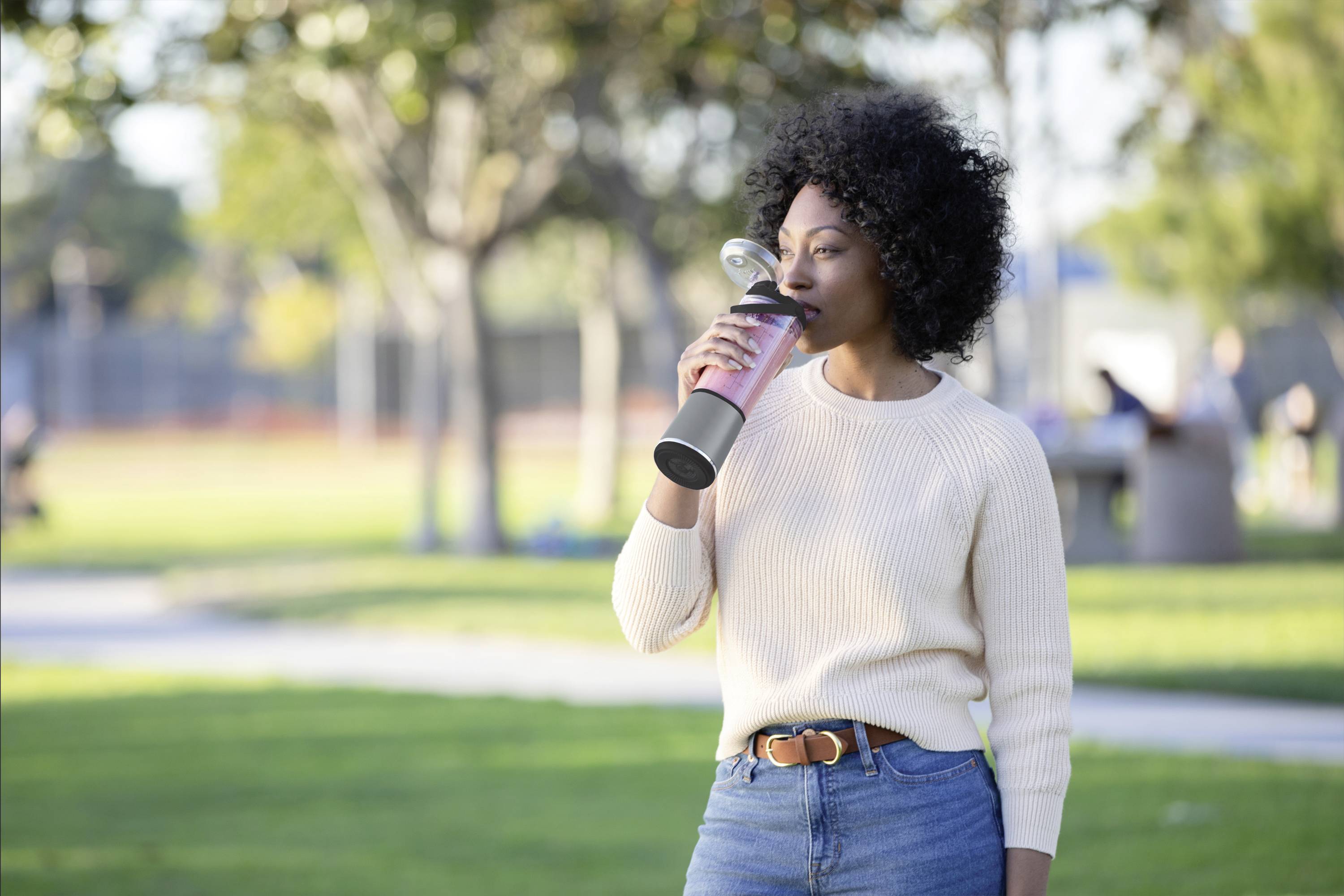 A woman is standing outdoors on a grassy area, drinking from a bottle, on a sunny day with trees in the background.