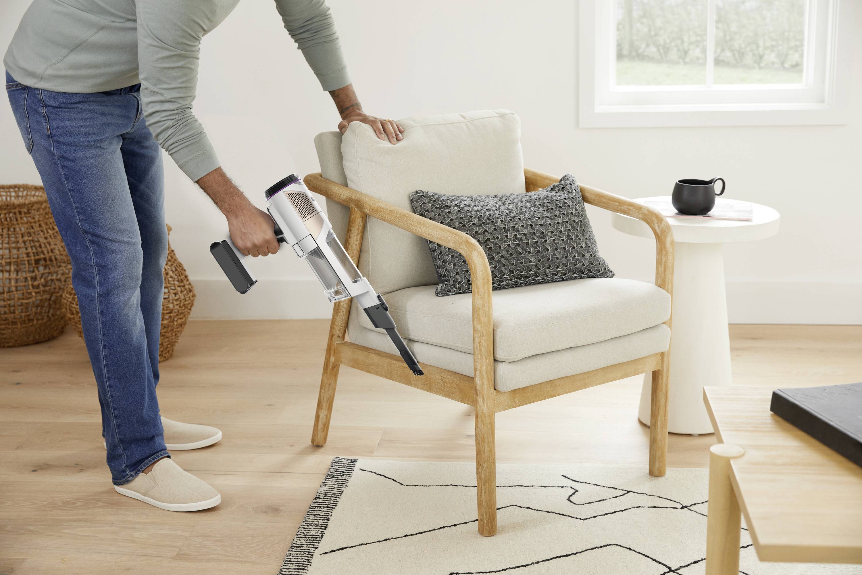 A person is hoovering a beige armchair in a modernly furnished room, with a small table, a cup, and a cushion.