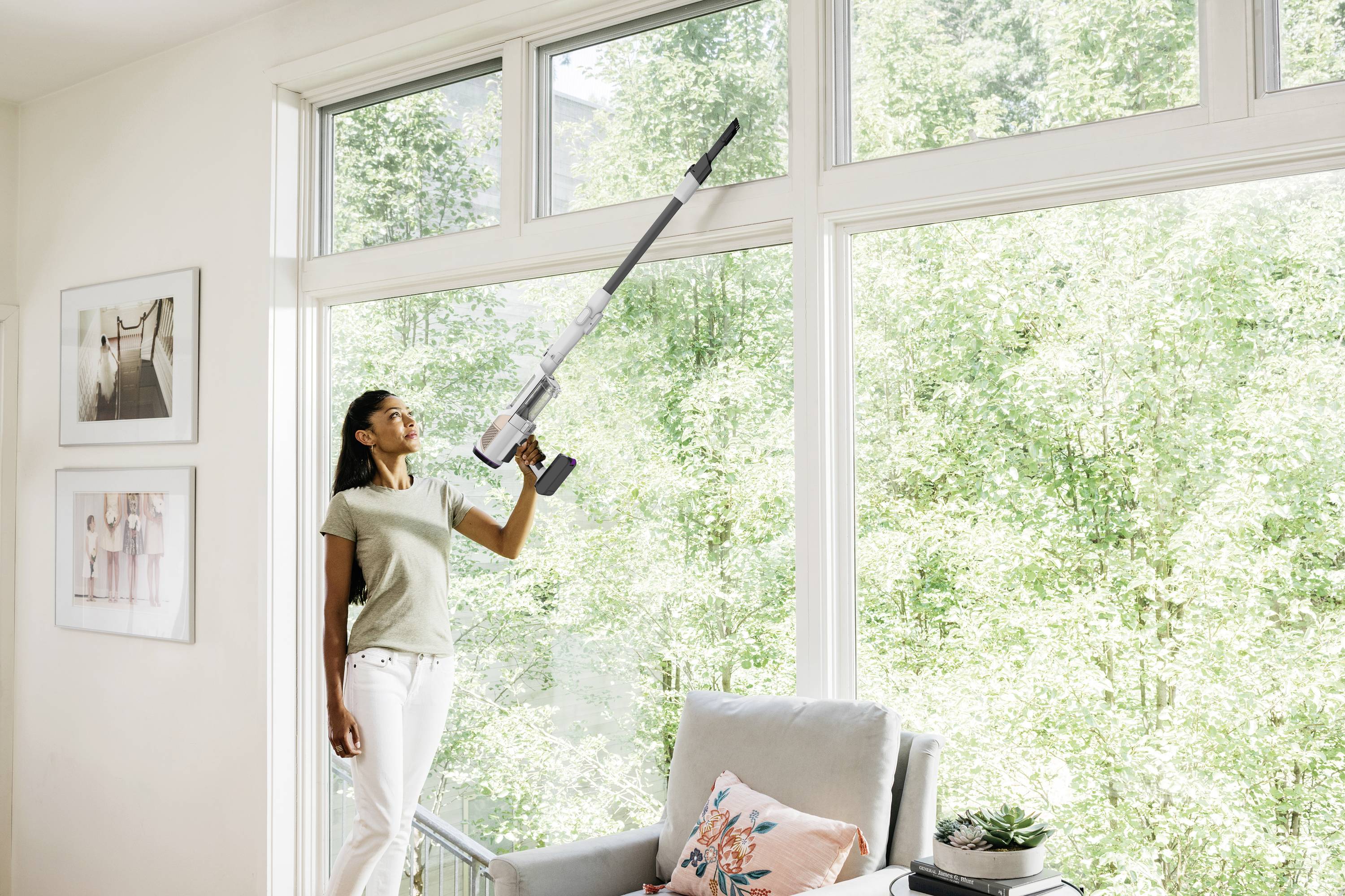 A woman is cleaning high windows with a cordless vacuum cleaner in a sunlit room with a view of the forest.