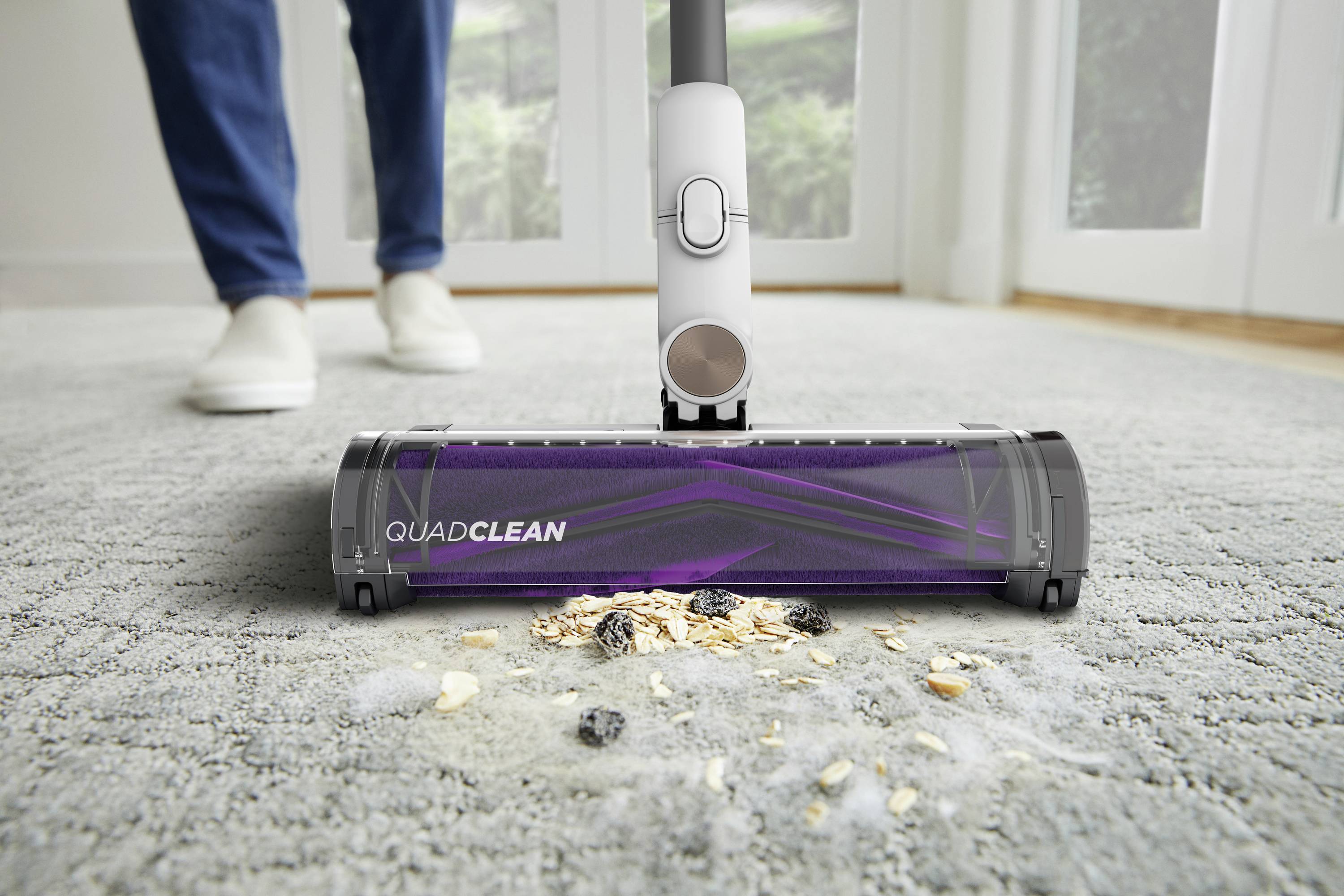 A purple vacuum cleaner is sucking up dirt and crumbs from a grey carpet. In the background, blurred legs of a person can be seen.