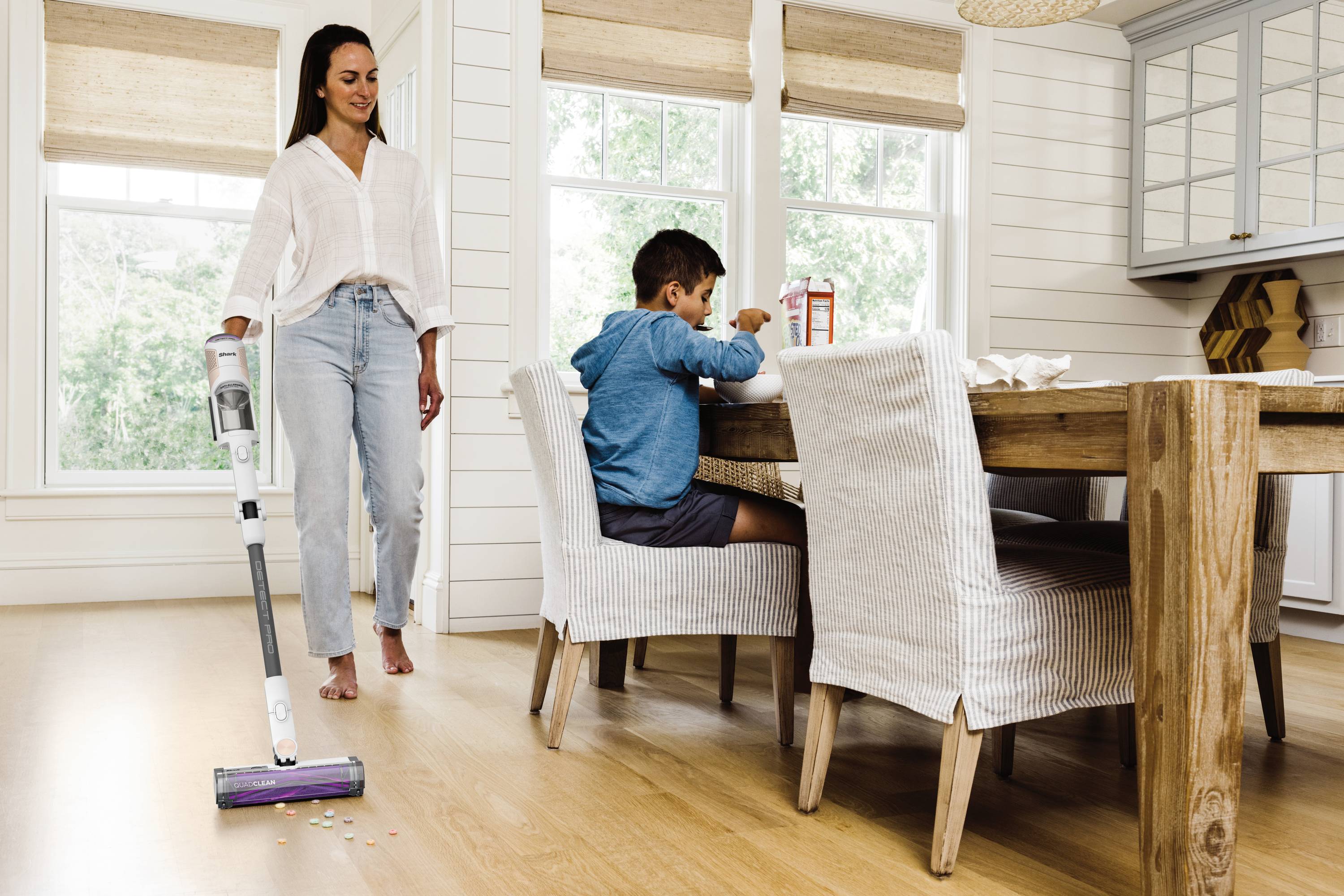 A woman is hoovering the floor in the dining room while a child sits at the table eating. Bright, welcoming environment with large windows.