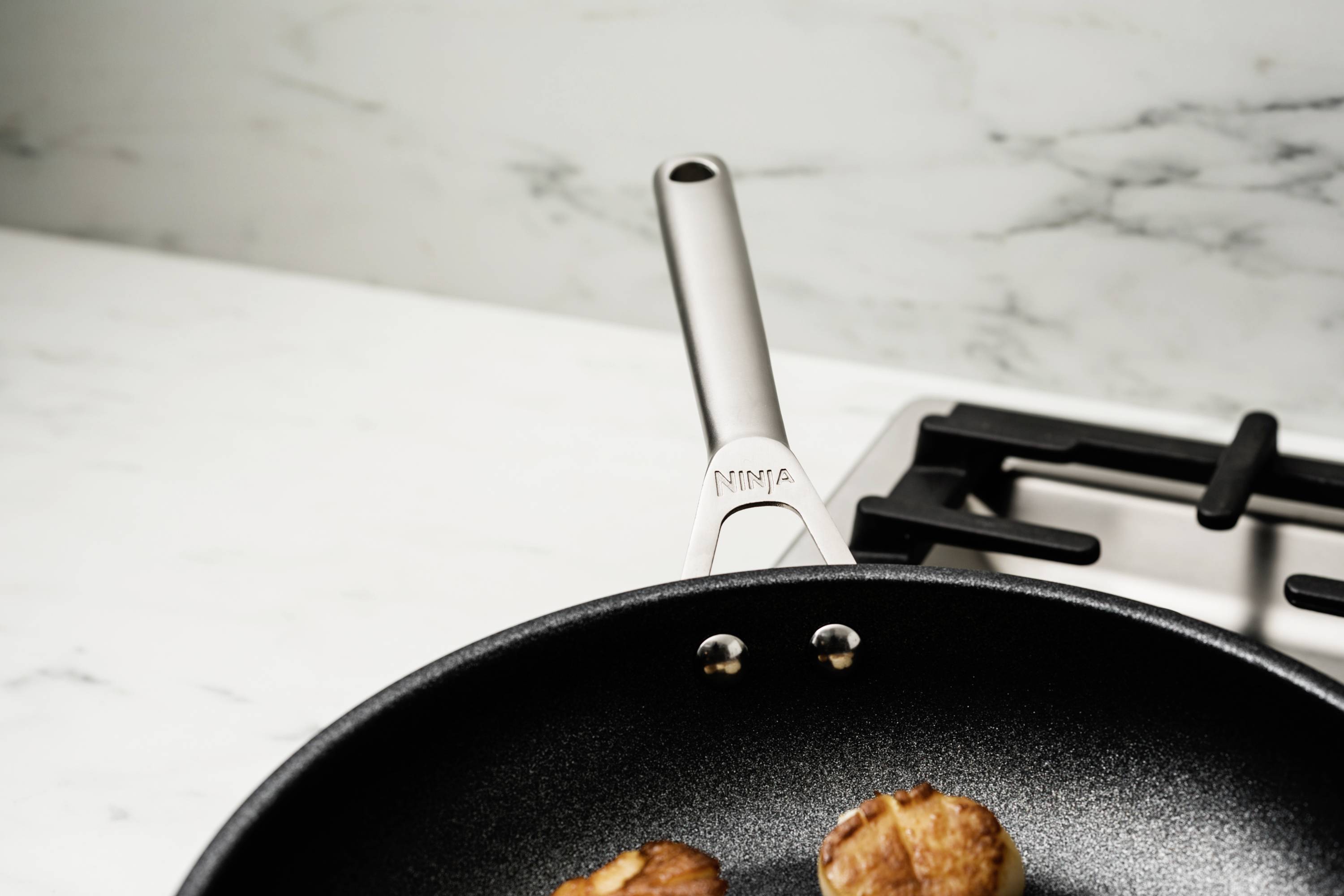 A black frying pan with fried mushrooms on a gas hob in a modern kitchen. The pan handle displays the brand name.