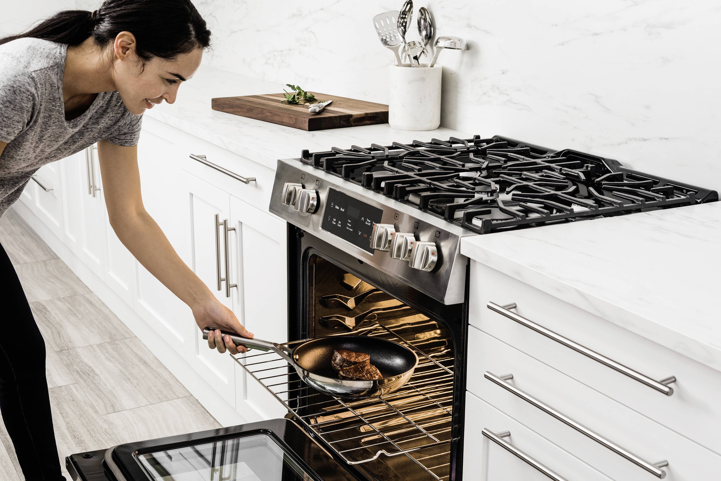 A person places a pan with fried meat into the oven. The kitchen is modern and clean, with white cupboards and a marble worktop.