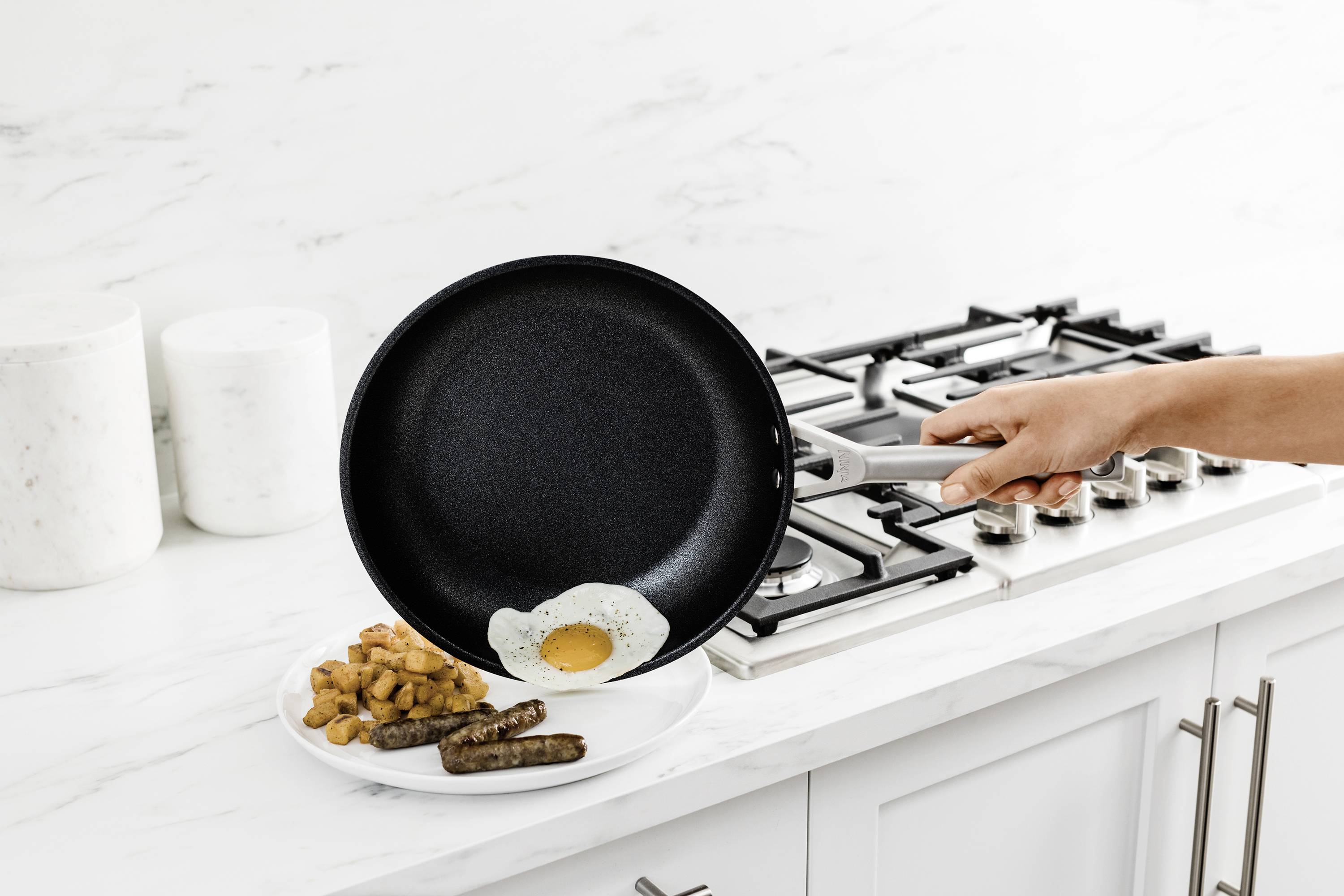 A hand tilts a pan, sliding a fried egg onto a plate of potatoes and sausages in a modern kitchen.