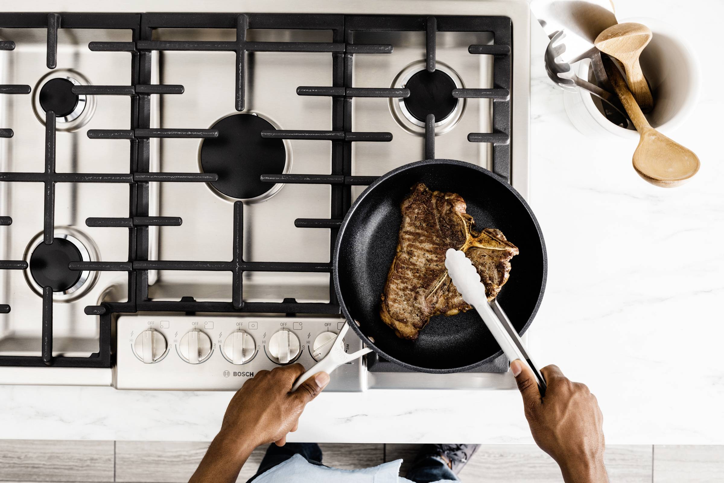 A person is frying a steak in a pan on a gas hob. Next to it is a container with wooden spoons.