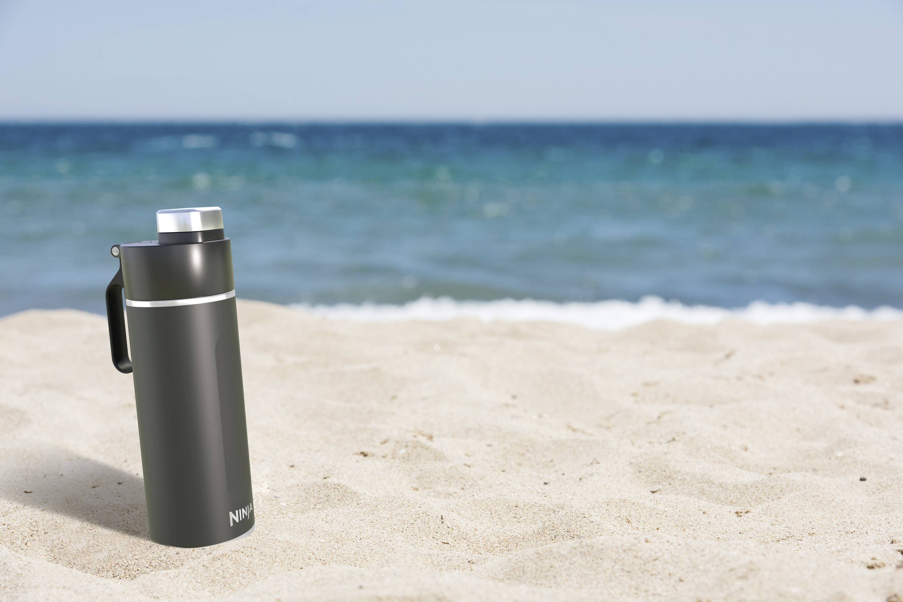 A black thermos flask sits on a sandy beach, overlooking the blue sea in the background.