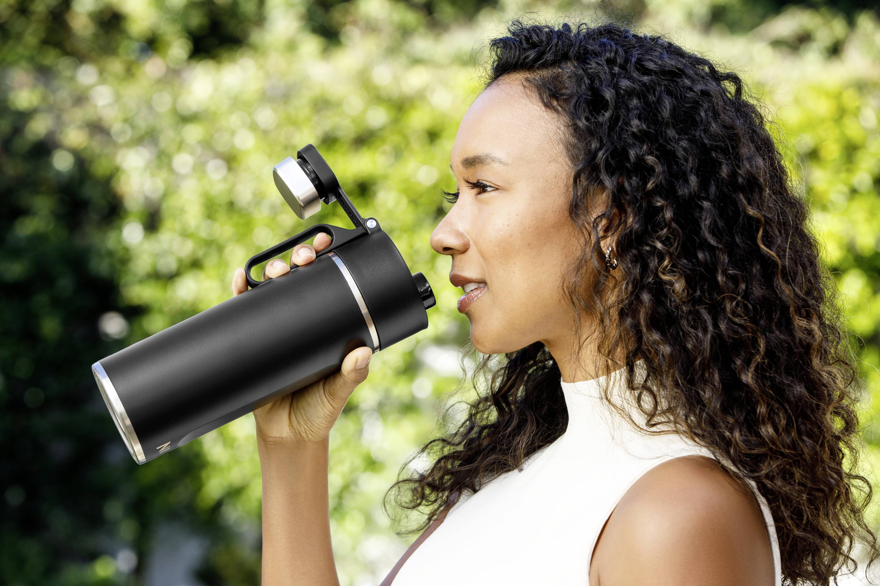 A woman is drinking from a black, reusable water bottle outdoors. Green trees can be seen in the background.