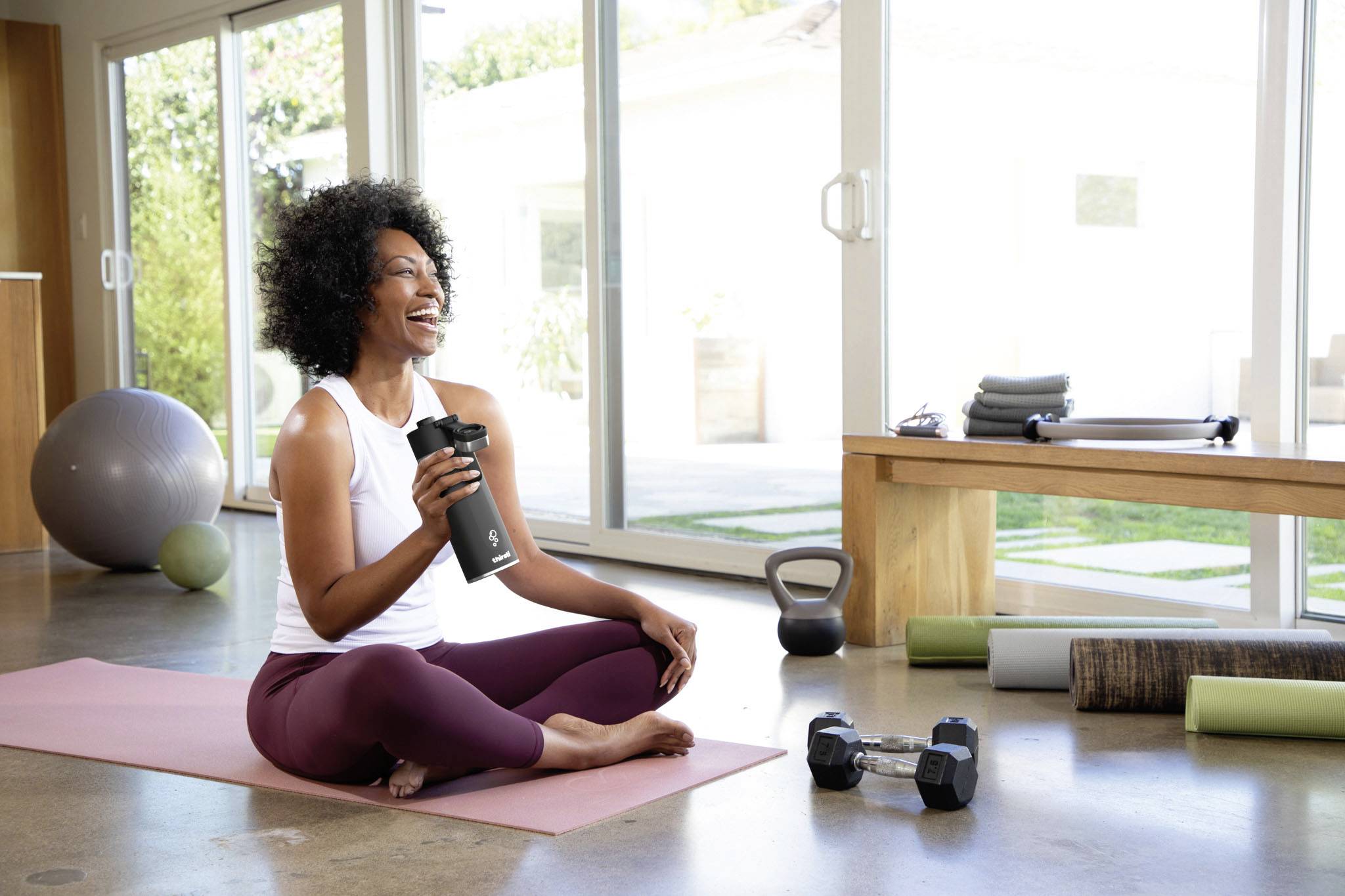 A woman sits on a yoga mat in a bright room, holding a water bottle and smiling. Weights and an exercise ball are visible in the background.