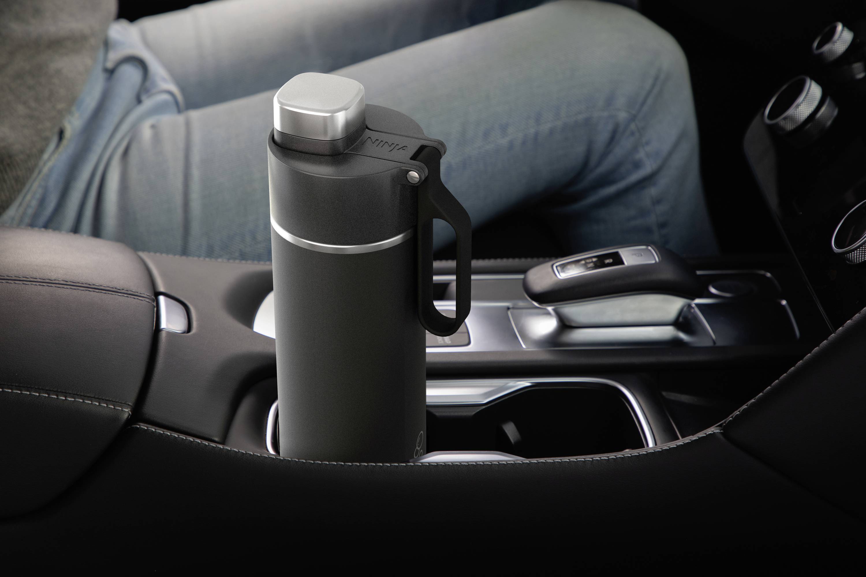 The image shows a cup holder in a car holding a grey thermos flask. A person is seated in the driver's seat.
