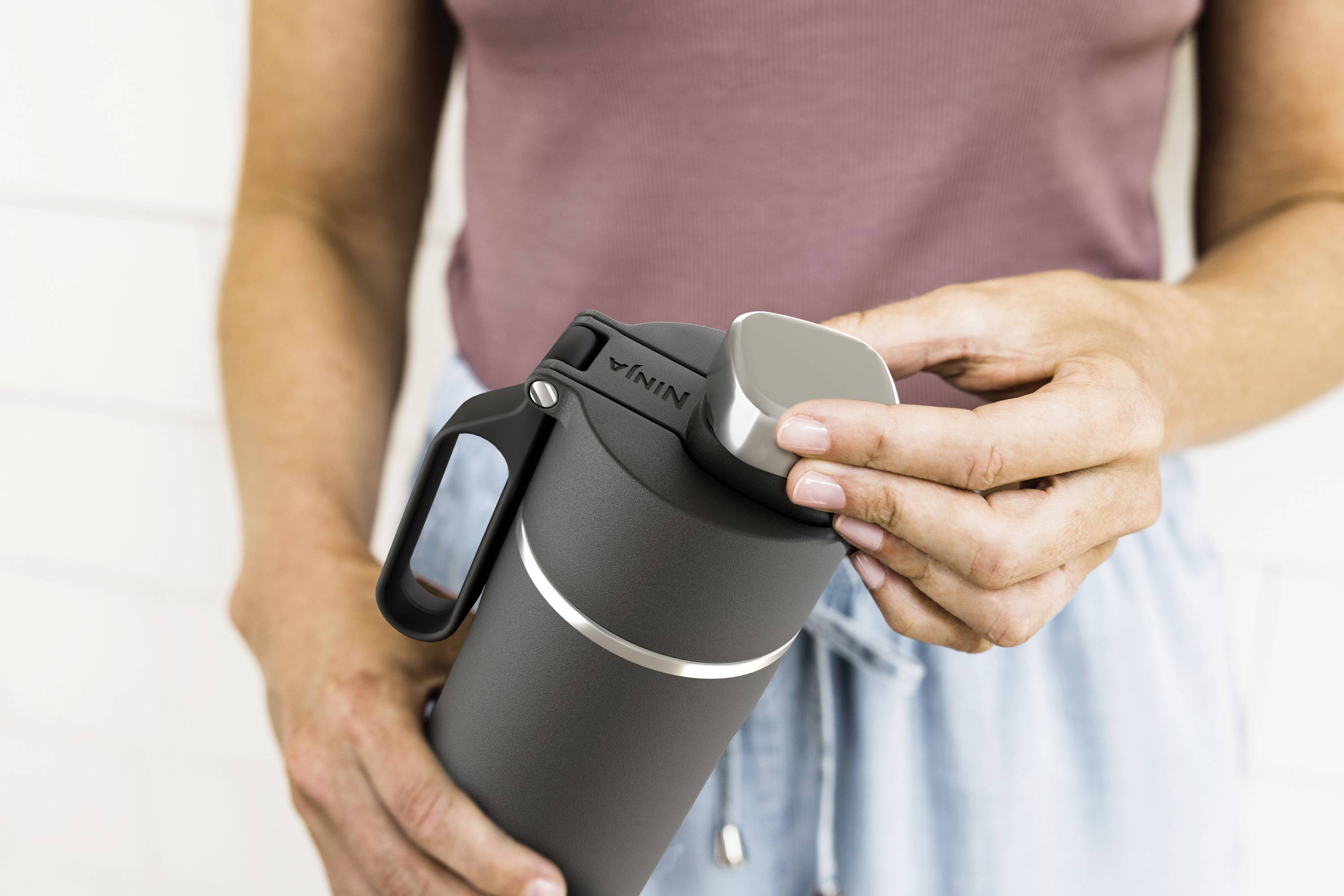 A person is holding a black thermos flask and unscrewing the lid. They are wearing a purple top and blue trousers.