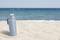 A blue thermos flask sits in the sand on the beach. Waves of the sea and a clear sky can be seen in the background.
