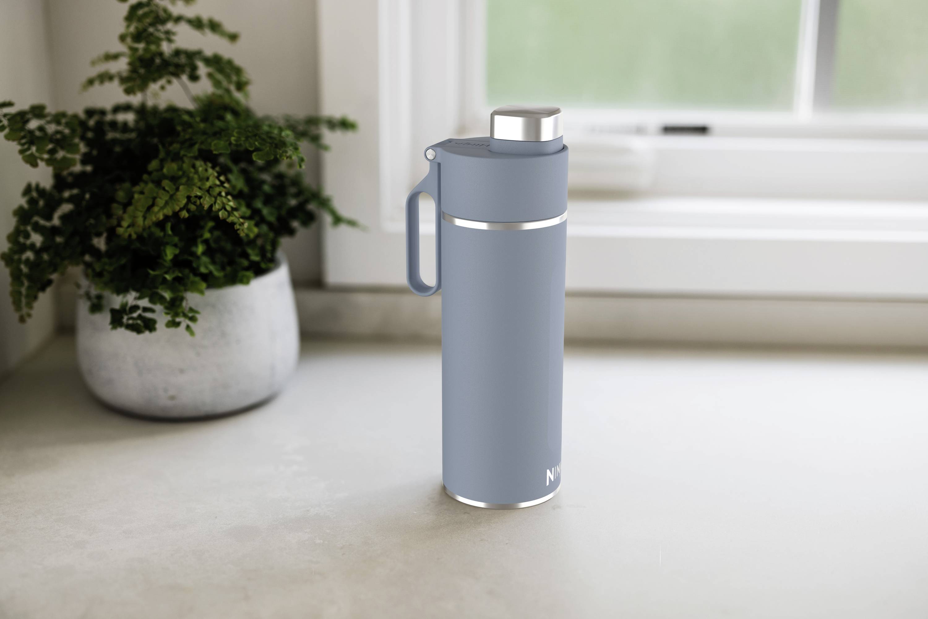 Grey thermos flask on kitchen worktop beside potted plant in front of window.