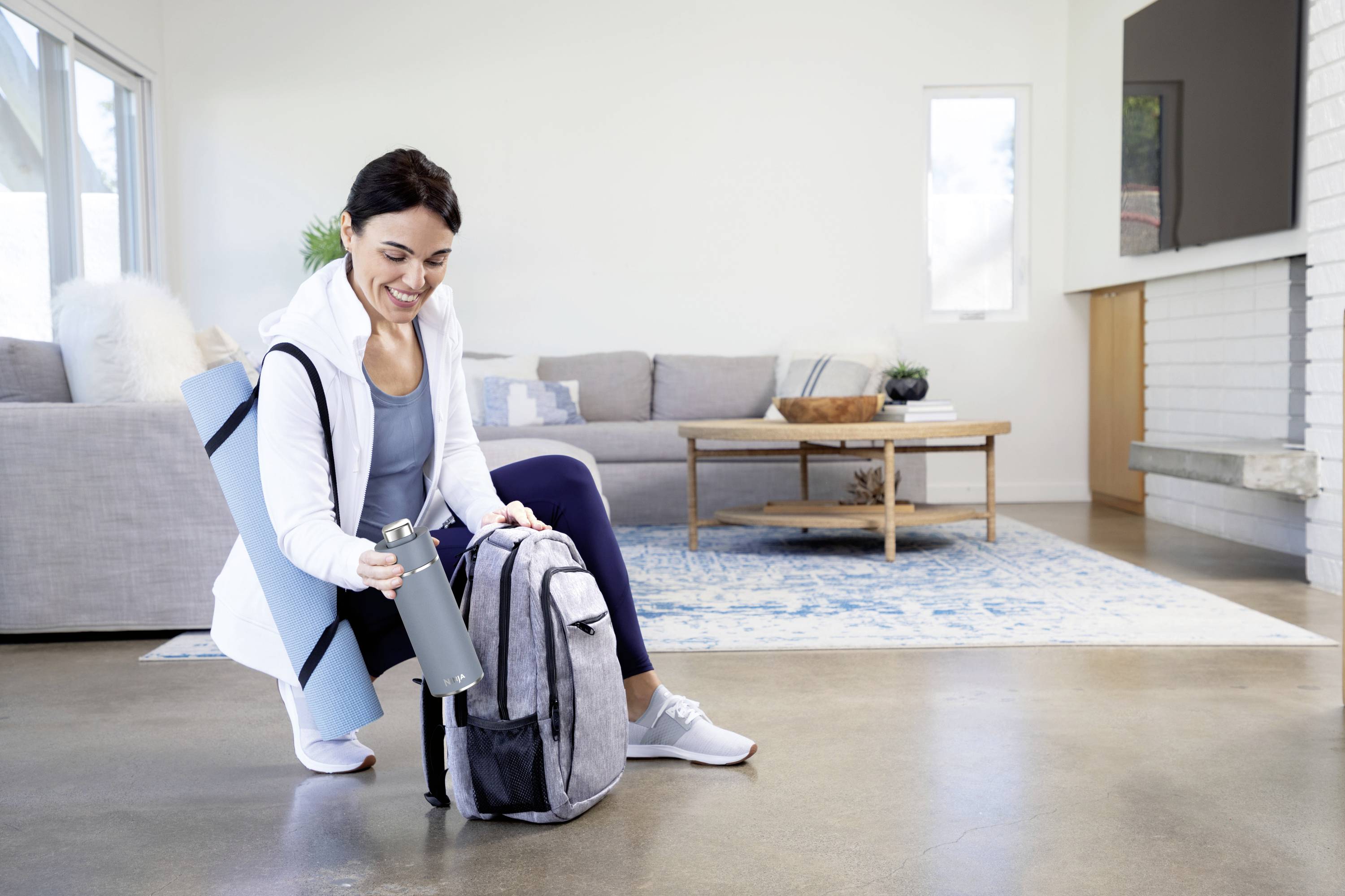 A woman in sportswear is sitting on the floor, placing a grey water bottle into her rucksack. A yoga mat is slung over her shoulder.