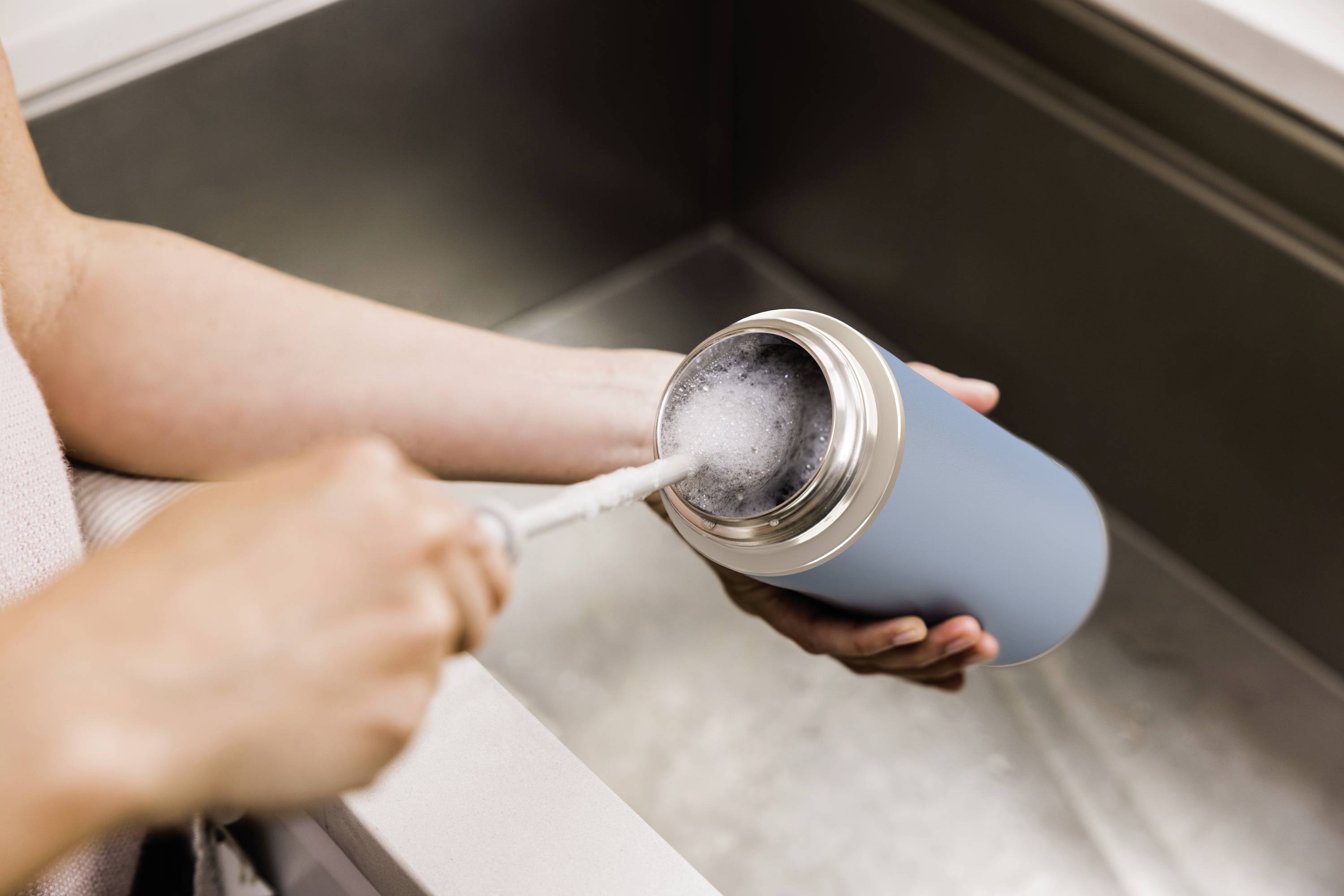 A person is cleaning a blue thermos flask at the sink with a brush. Water is being used to rinse it.