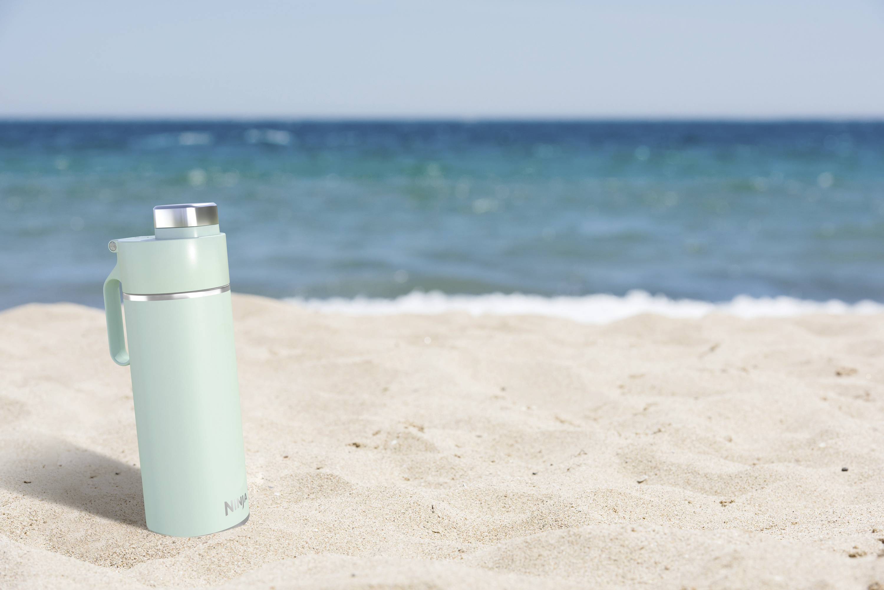 A green thermos flask sits on light sand in front of a blurred, blue ocean and sky in the background.