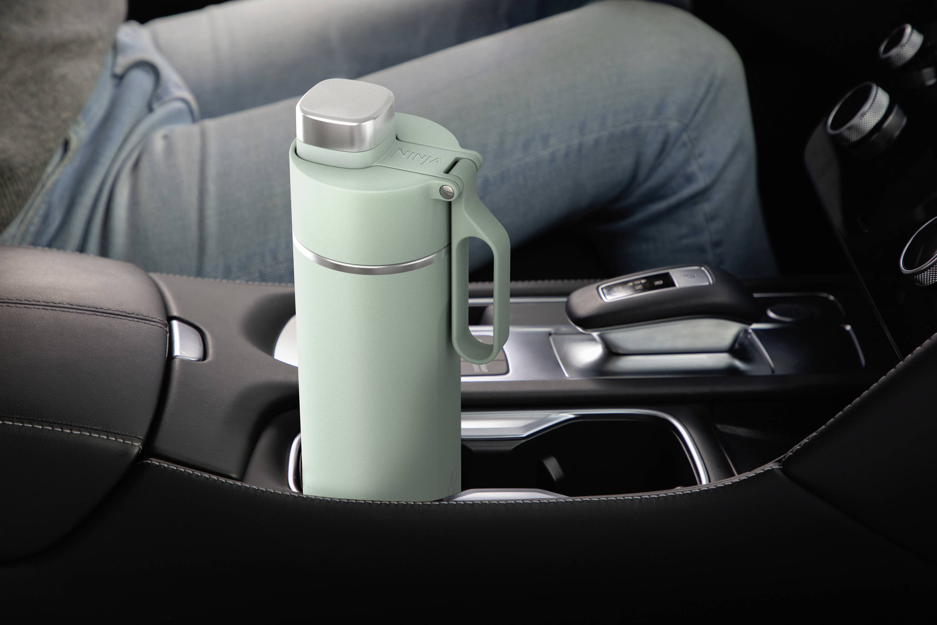 A person is sitting in a car in the passenger seat. A mint green thermos flask sits in the centre console.