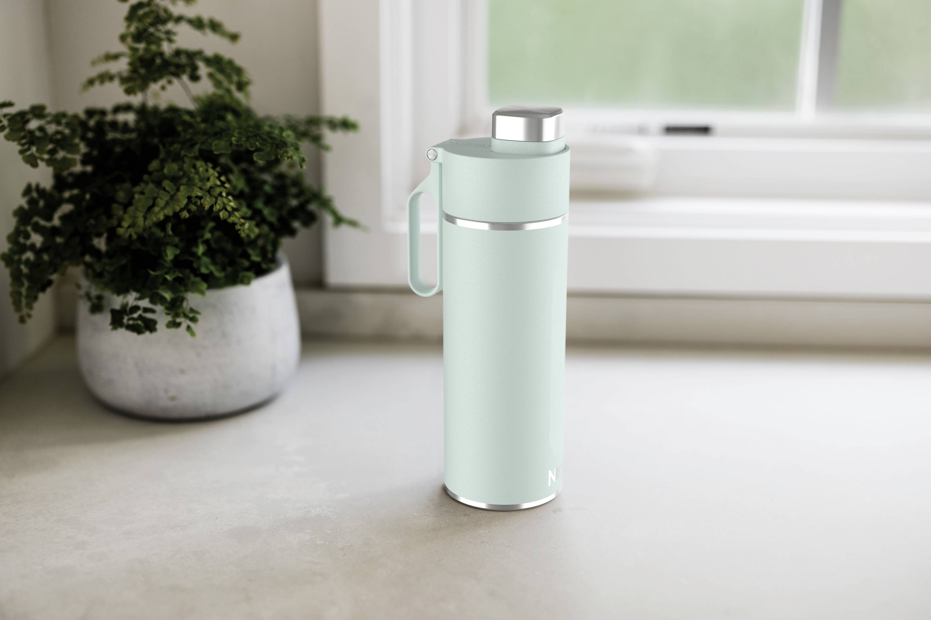 A mint green thermos flask sits on a kitchen worktop next to a plant in front of a window.