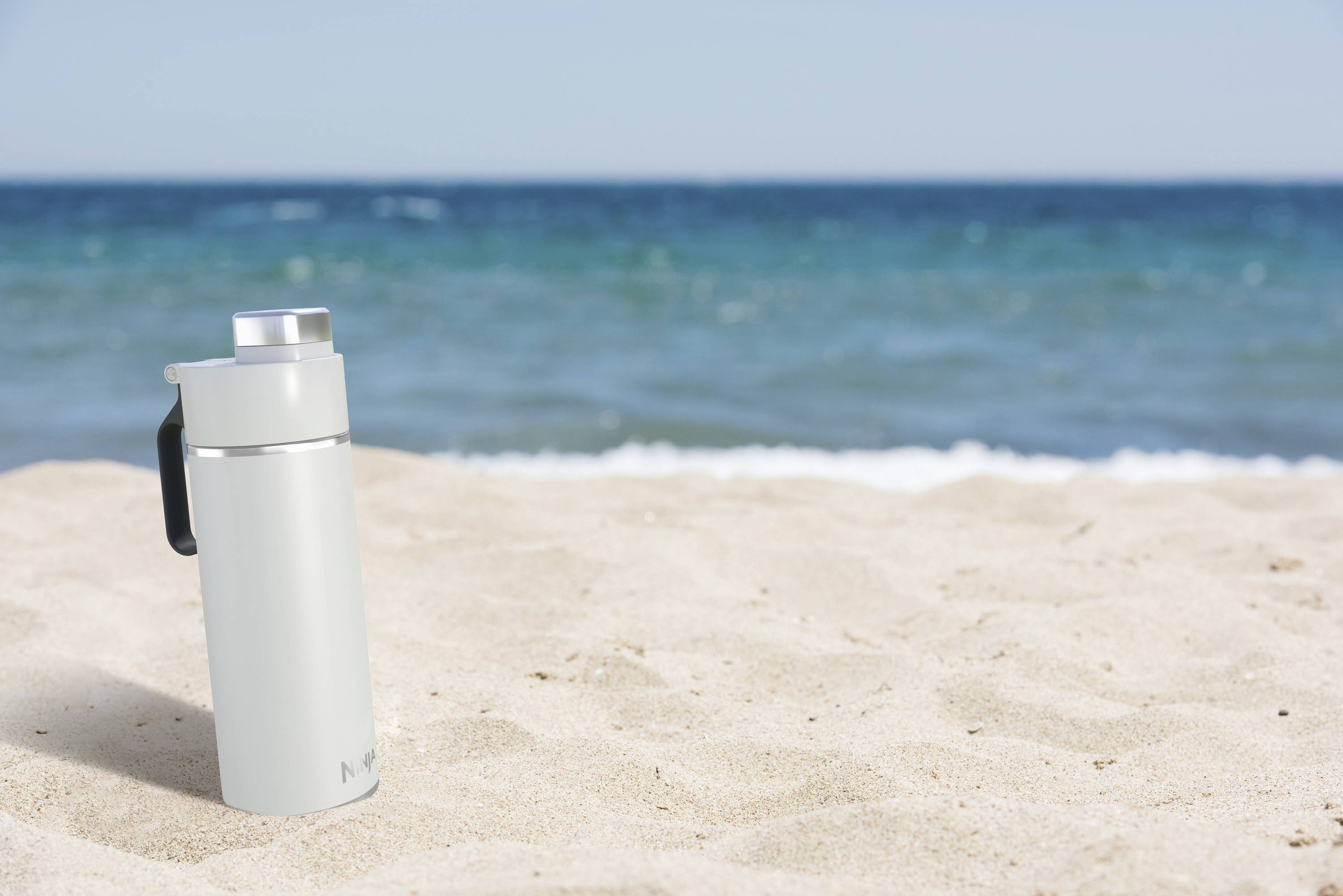 A silver thermal mug sits on light sand in front of a blue sea. The sky is clear.