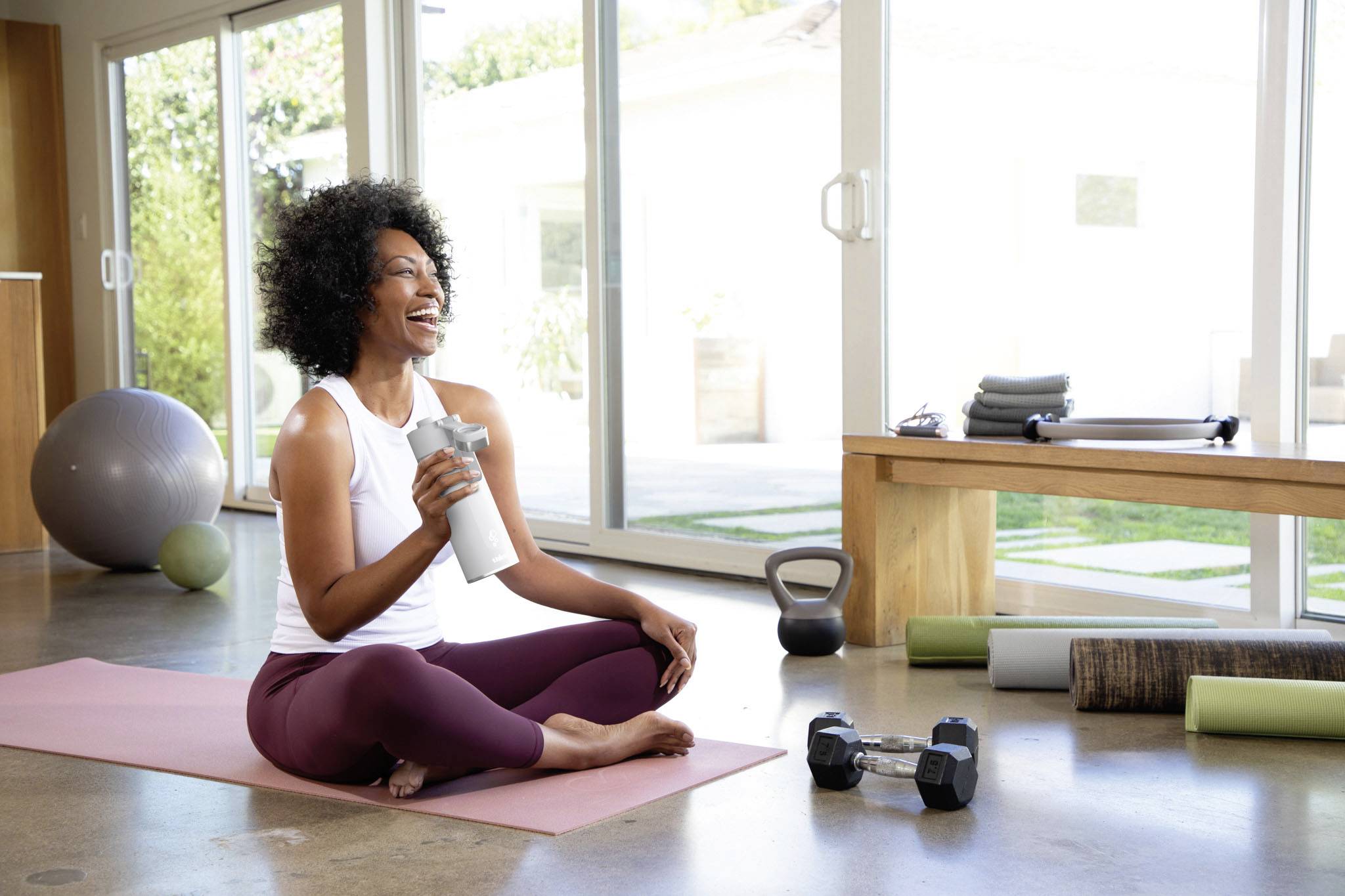 A woman sits smiling on a yoga mat, holding a water bottle. Dumbbells and fitness equipment are beside her. Bright, airy environment.