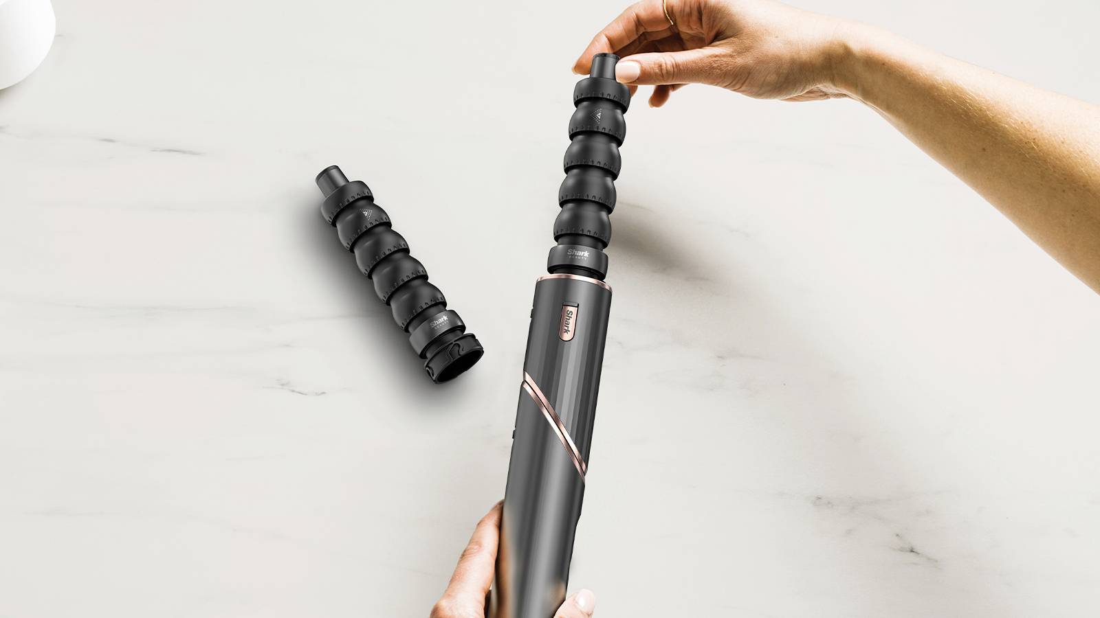 A hand is changing a detachable curling attachment on a hair styling tool. An additional attachment lies beside it.