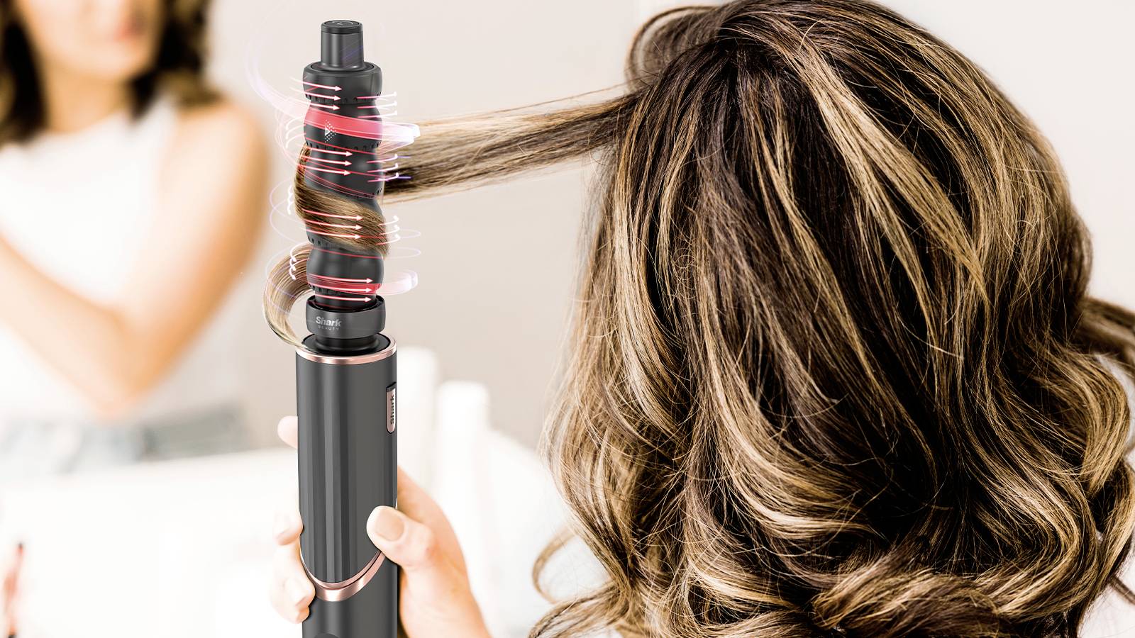 A woman curls her hair with an automatic curling wand in the bathroom. In the background, her blurred reflection is mirrored in the mirror.