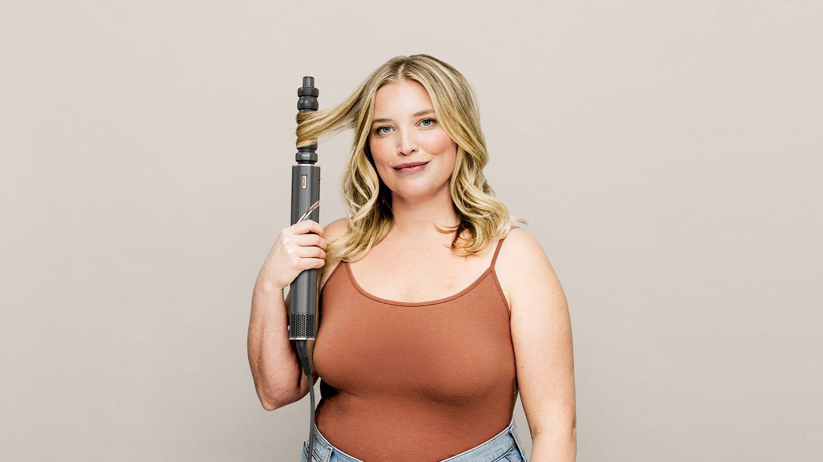 A person with blonde hair is holding a curling iron, wearing a brown top and jeans, smiling warmly at the camera.