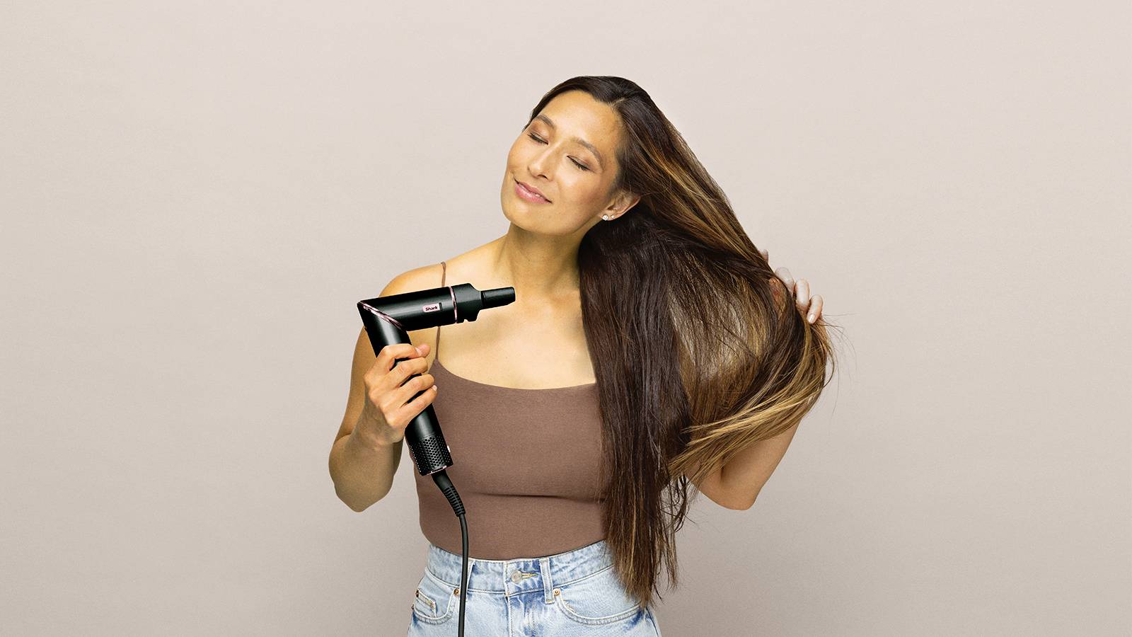 A woman with long hair is using a hairdryer. She appears relaxed and focused on styling her hair.