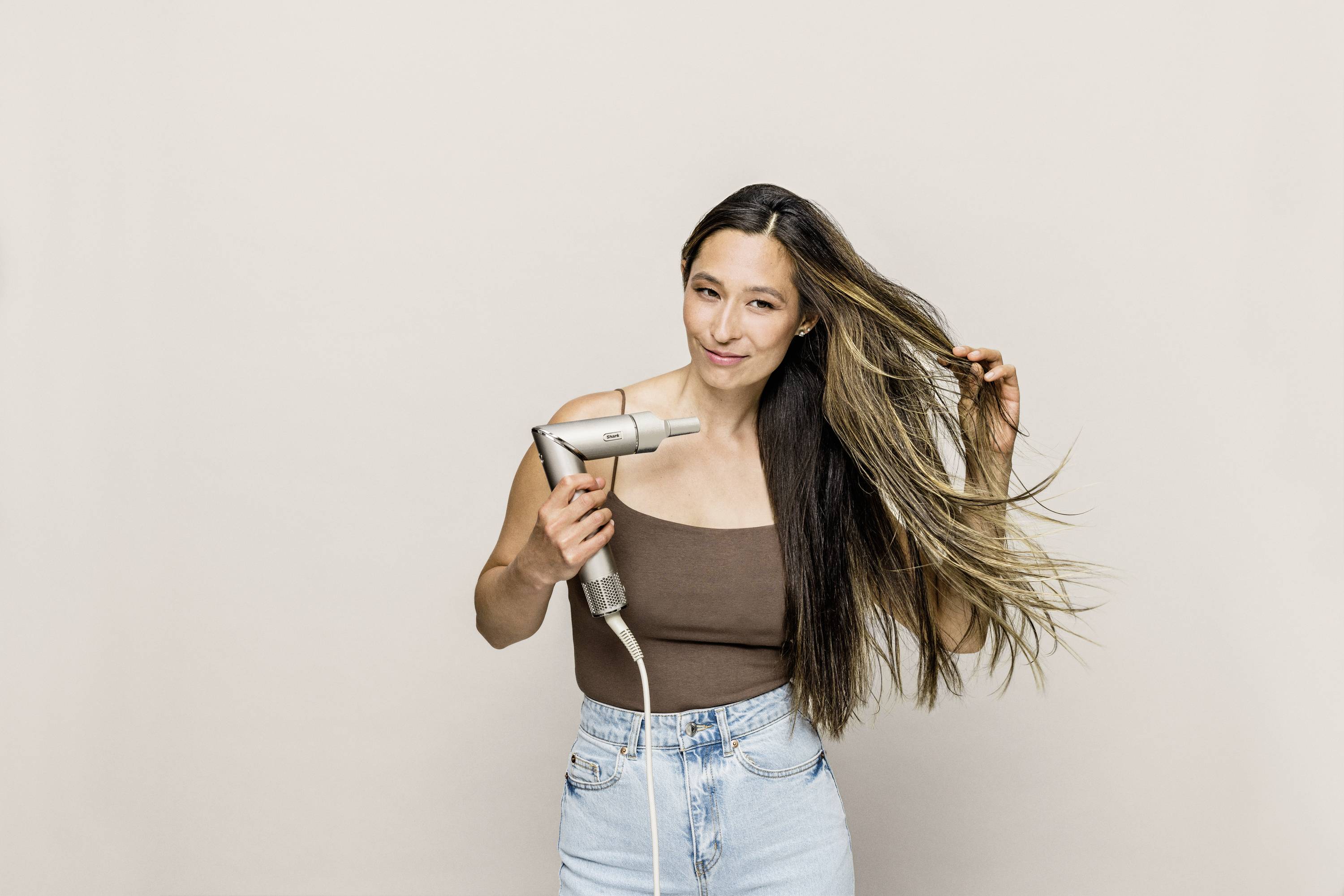 A woman is drying her long, brunette hair with a hairdryer. She is wearing a brown top and jeans against a neutral background.
