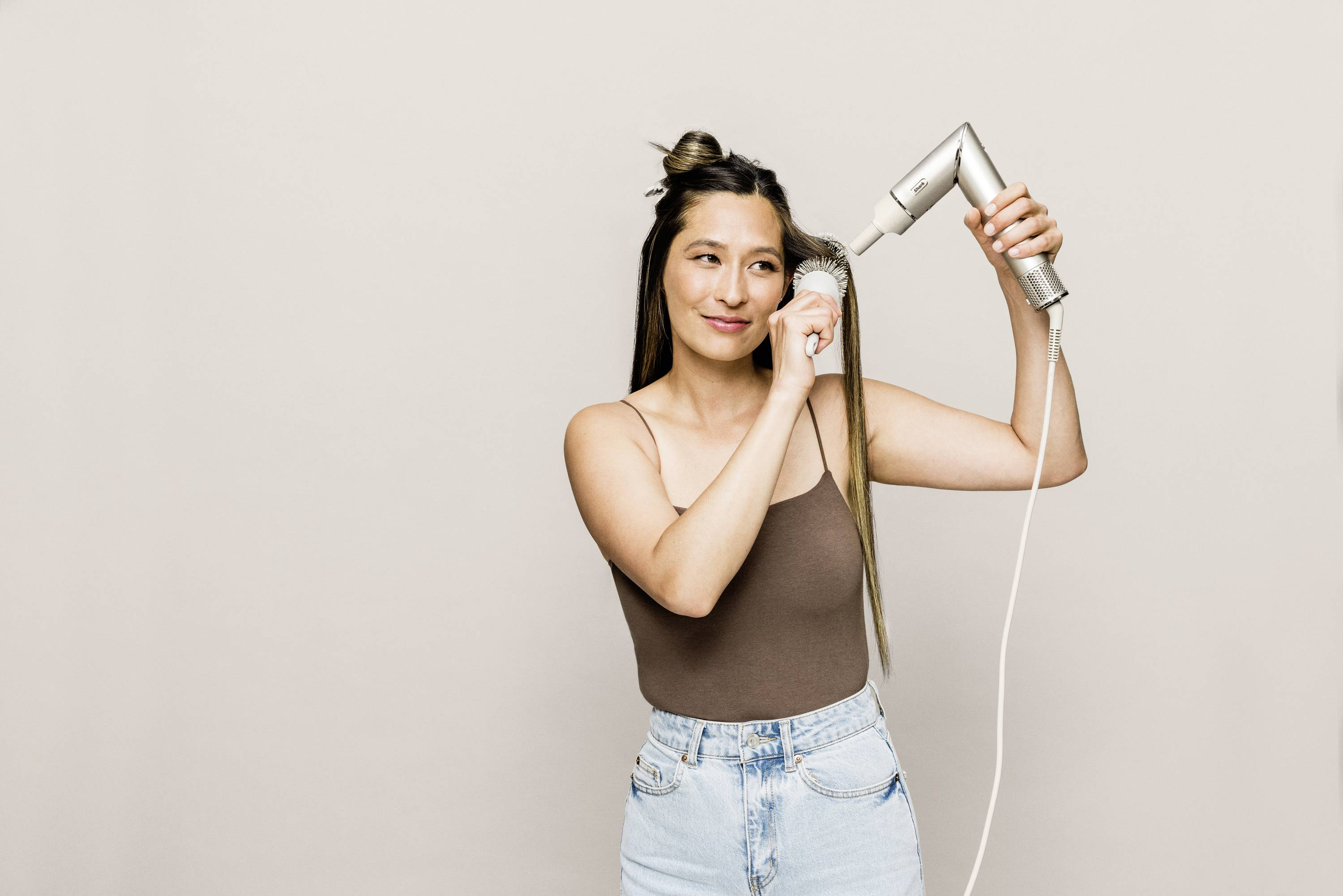 A woman is drying her long hair with a hairdryer, wearing a brown top and jeans, with a slight smile, against a neutral background.