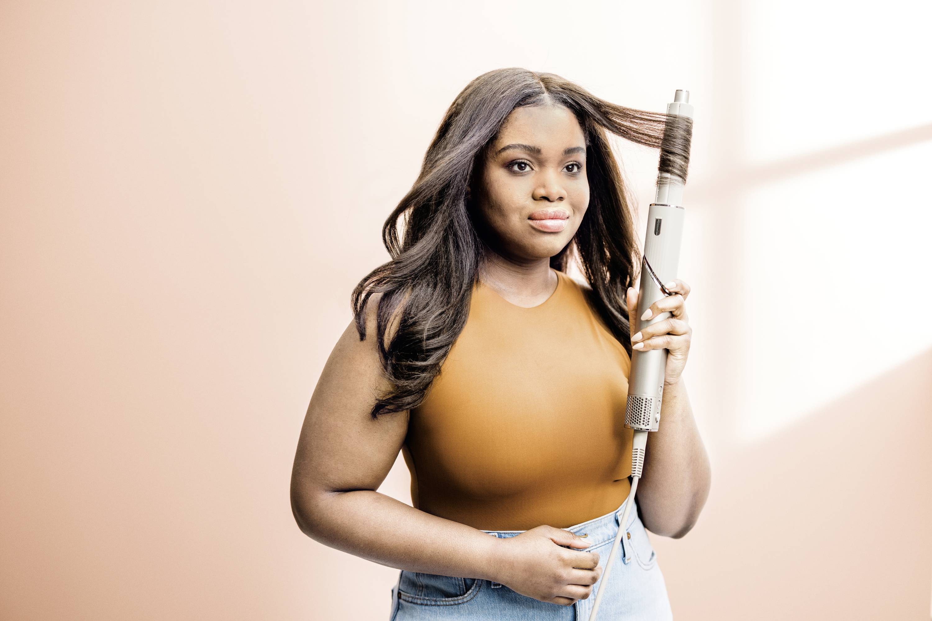 A woman with long hair is using a curling iron. She is wearing an orange top and jeans, with a light-coloured wall in the background.
