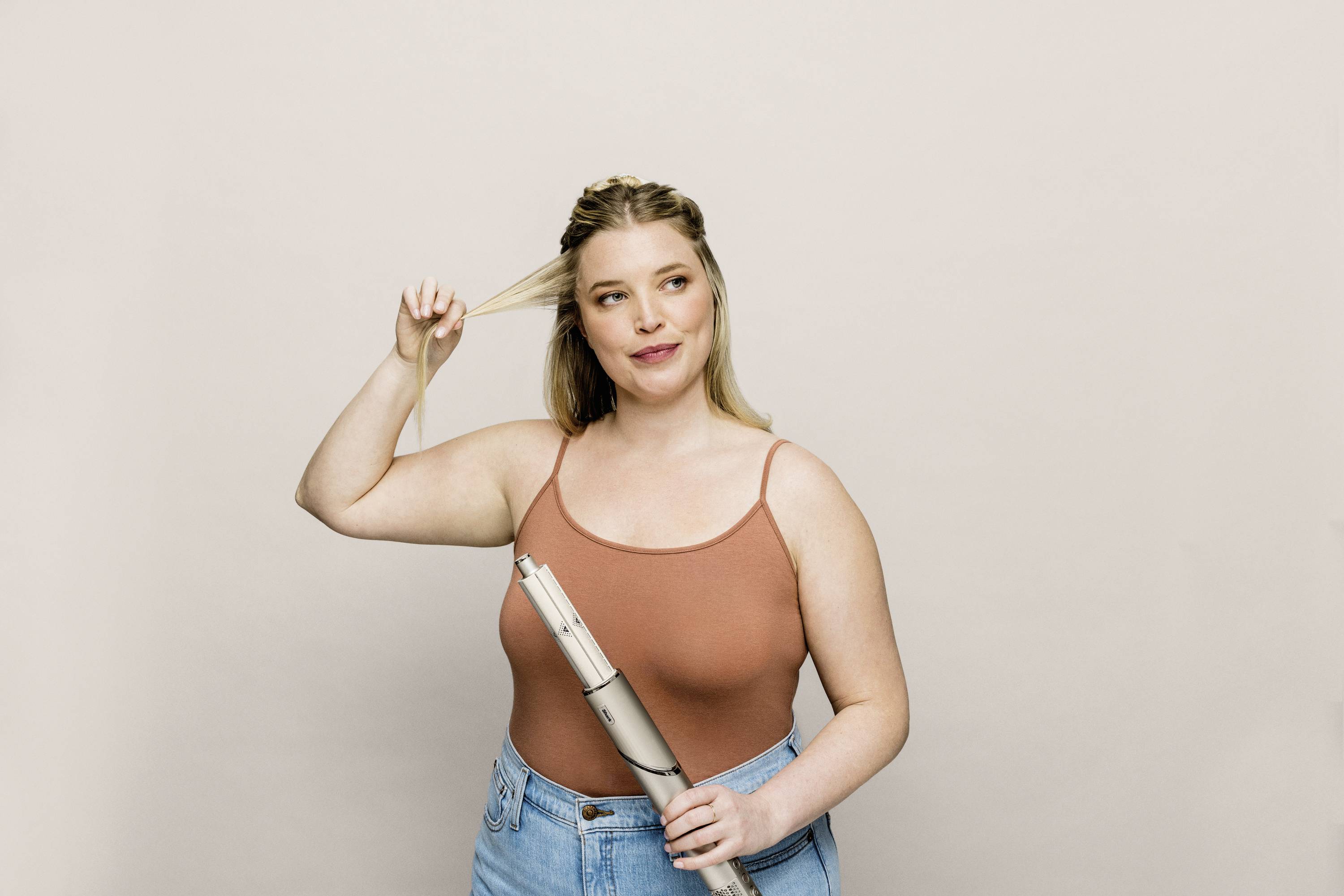 Woman with blonde hair holding a strand of hair and a curling iron. She is wearing a brown top and blue jeans against a neutral background.