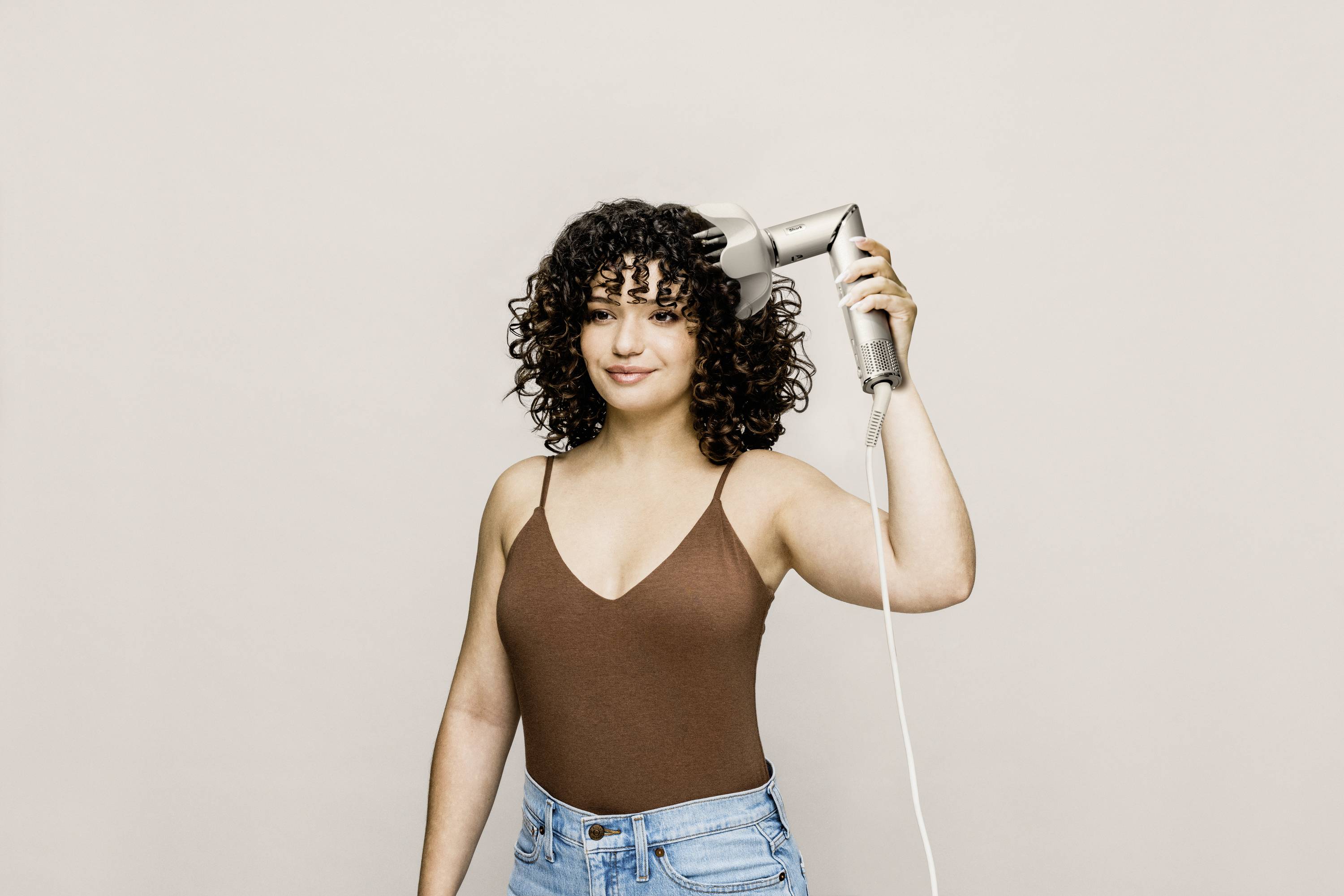 A person with curly hair is using a hairdryer. They are wearing a brown top and blue jeans, smiling and standing against a neutral background.
