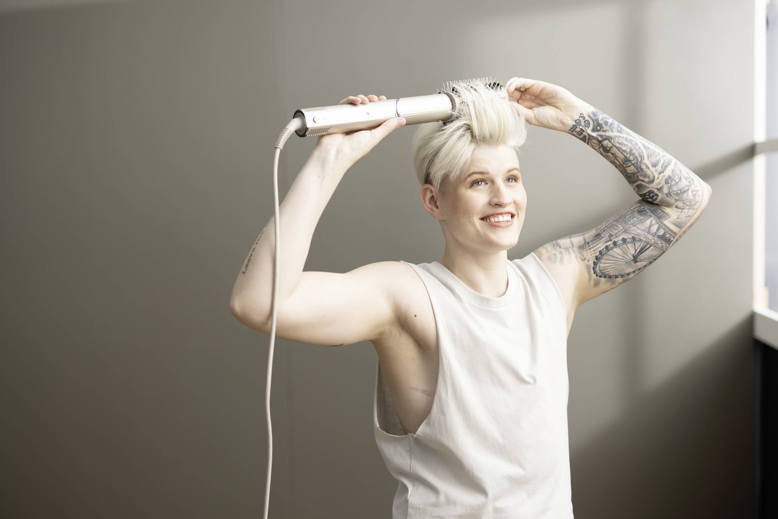 A person with tattoos is blow-drying their short, blonde hair with a round brush and smiling.