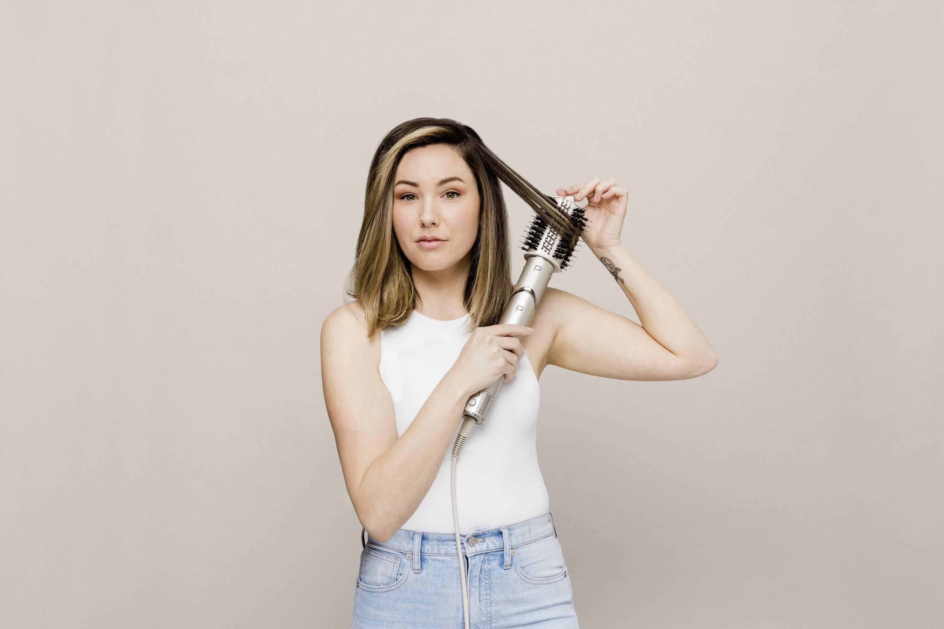 A woman styles her hair with a hot air brush against a neutral background. She is wearing a white top and blue jeans.