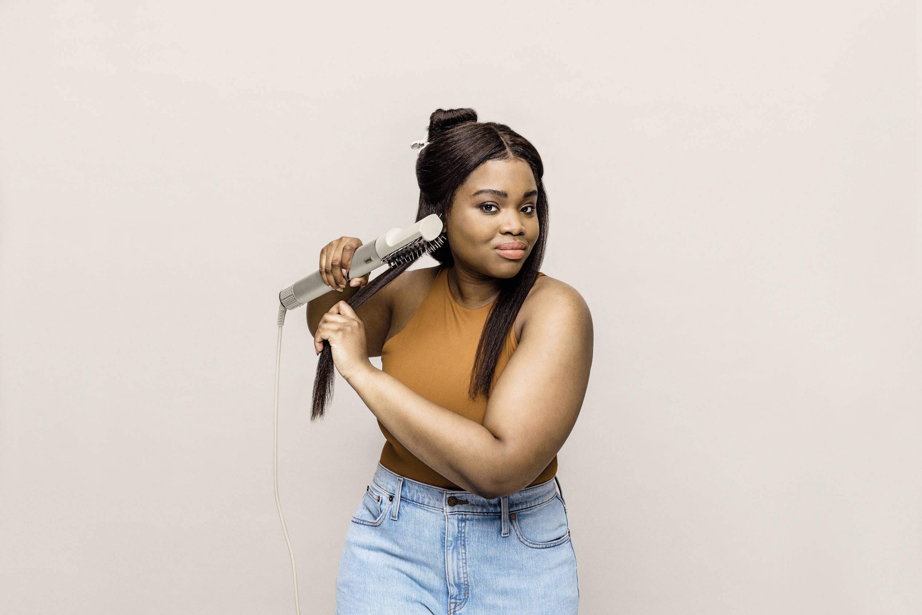 A woman with dark hair is straightening it with a hair straightener. She is wearing a sleeveless, brown top and blue jeans.