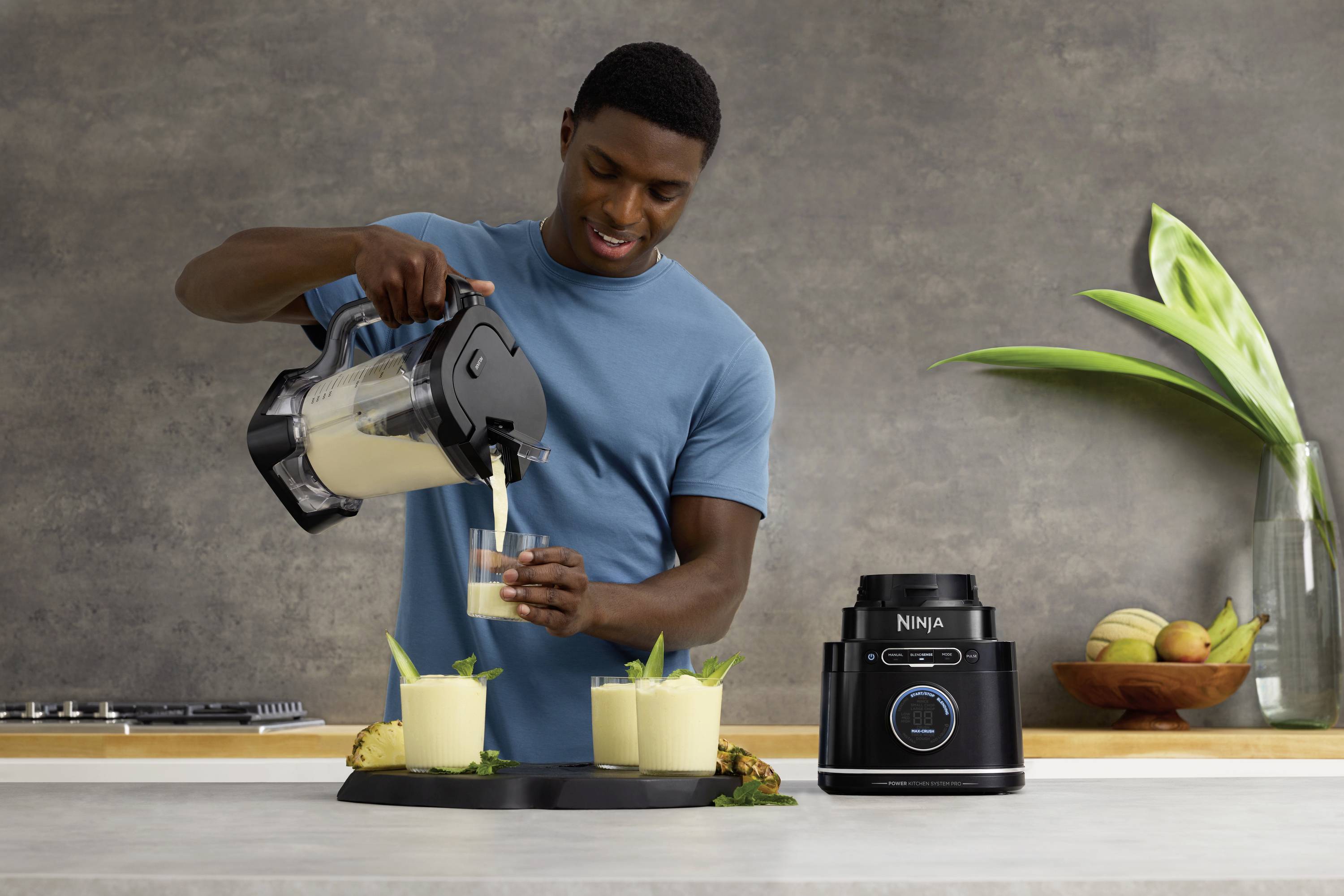 A person is pouring a smoothie from a blender jug into a glass. On the table, there are three filled glasses and a blender.