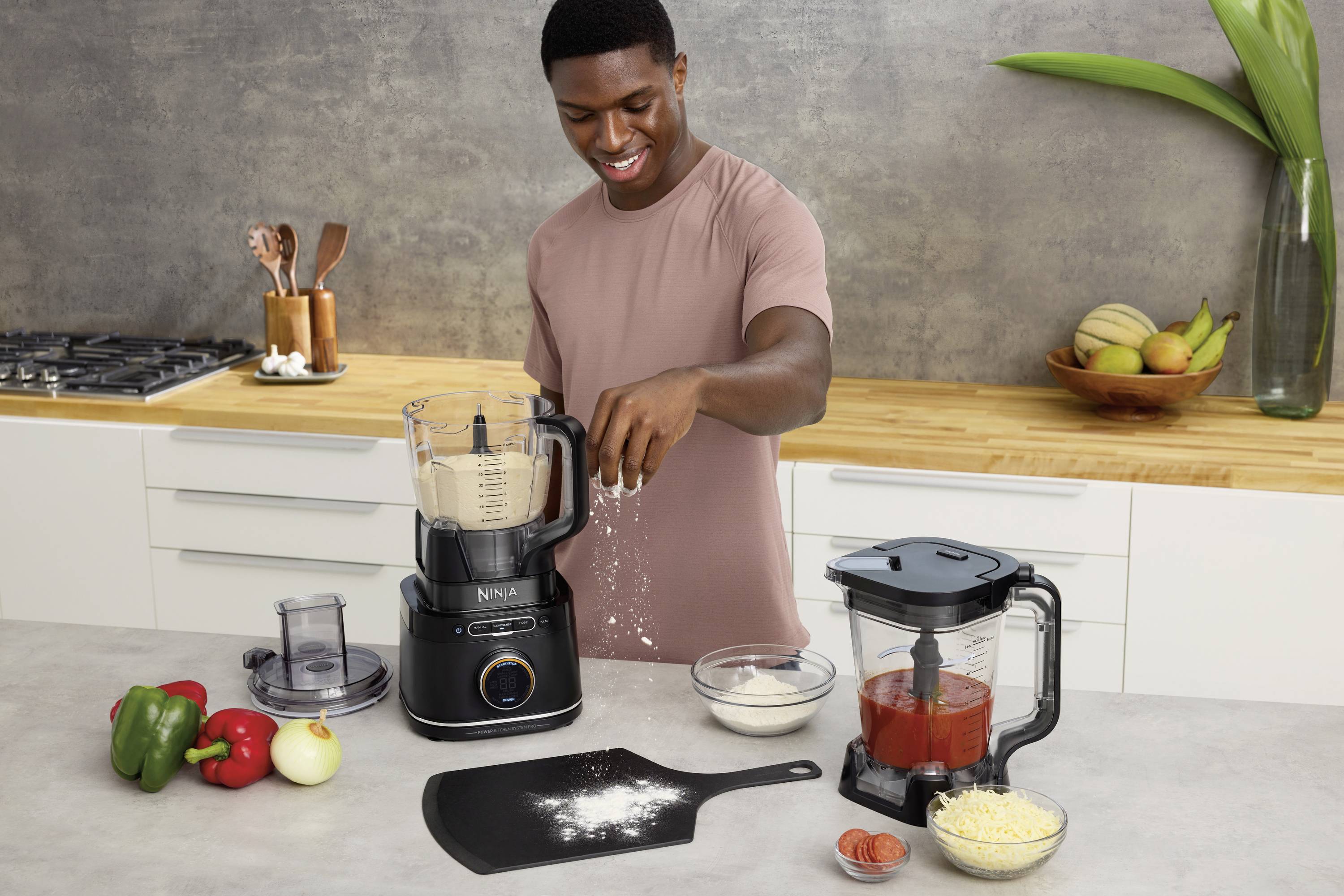 A man in a kitchen is sprinkling flour into a blender. On the worktop are peppers, an onion, cheese and a bowl of tomato sauce.