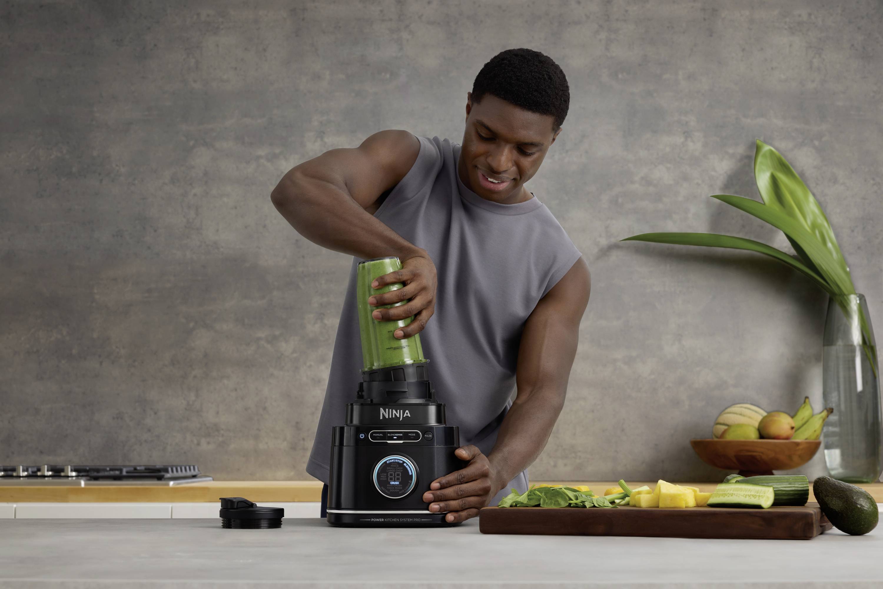 A person is preparing a green smoothie using a blender. Fruits and vegetables are laid out on the worktop.