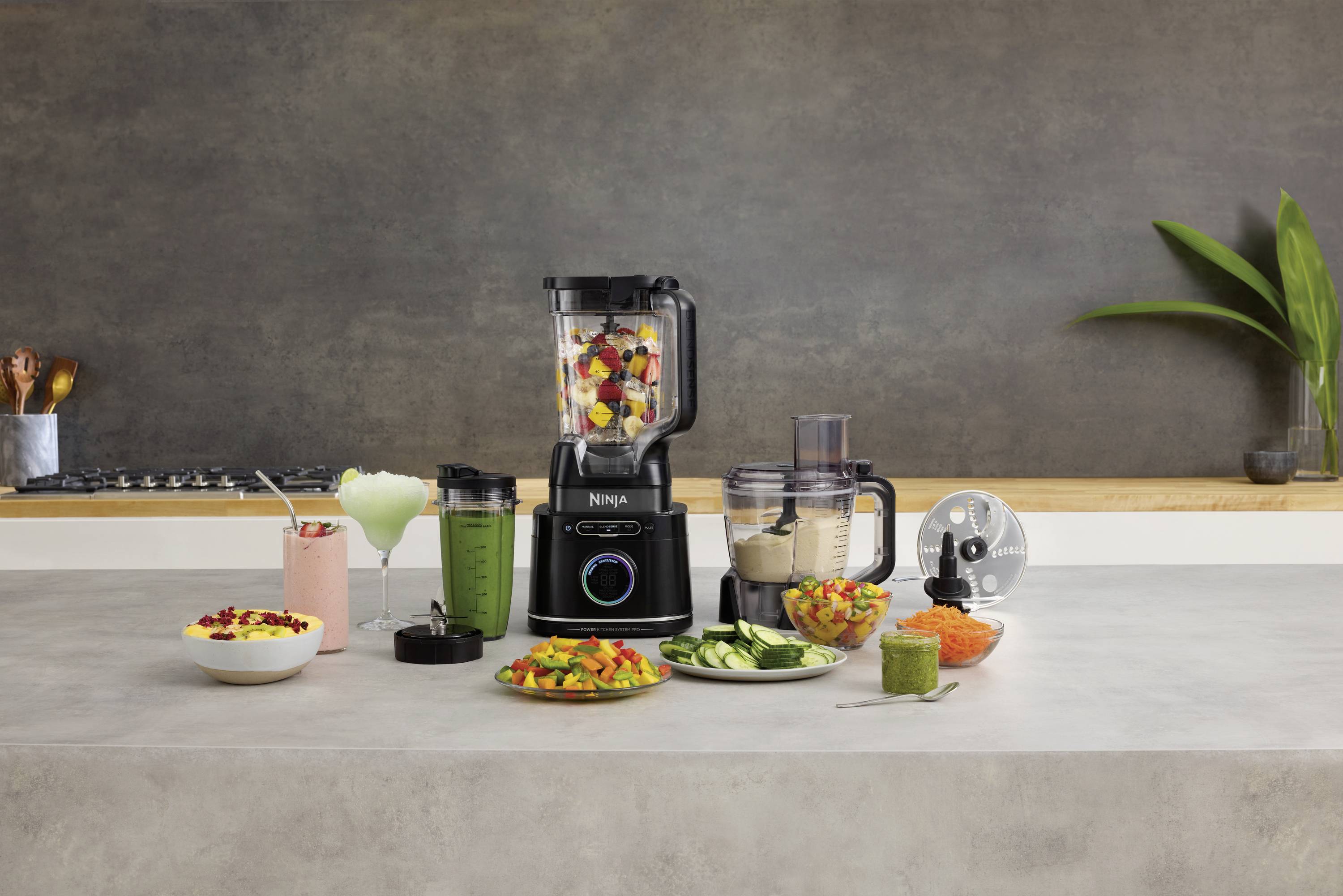 A kitchen appliance stands on a worktop, surrounded by chopped fruit, vegetables, and mixing containers. In the background, a modern kitchen.