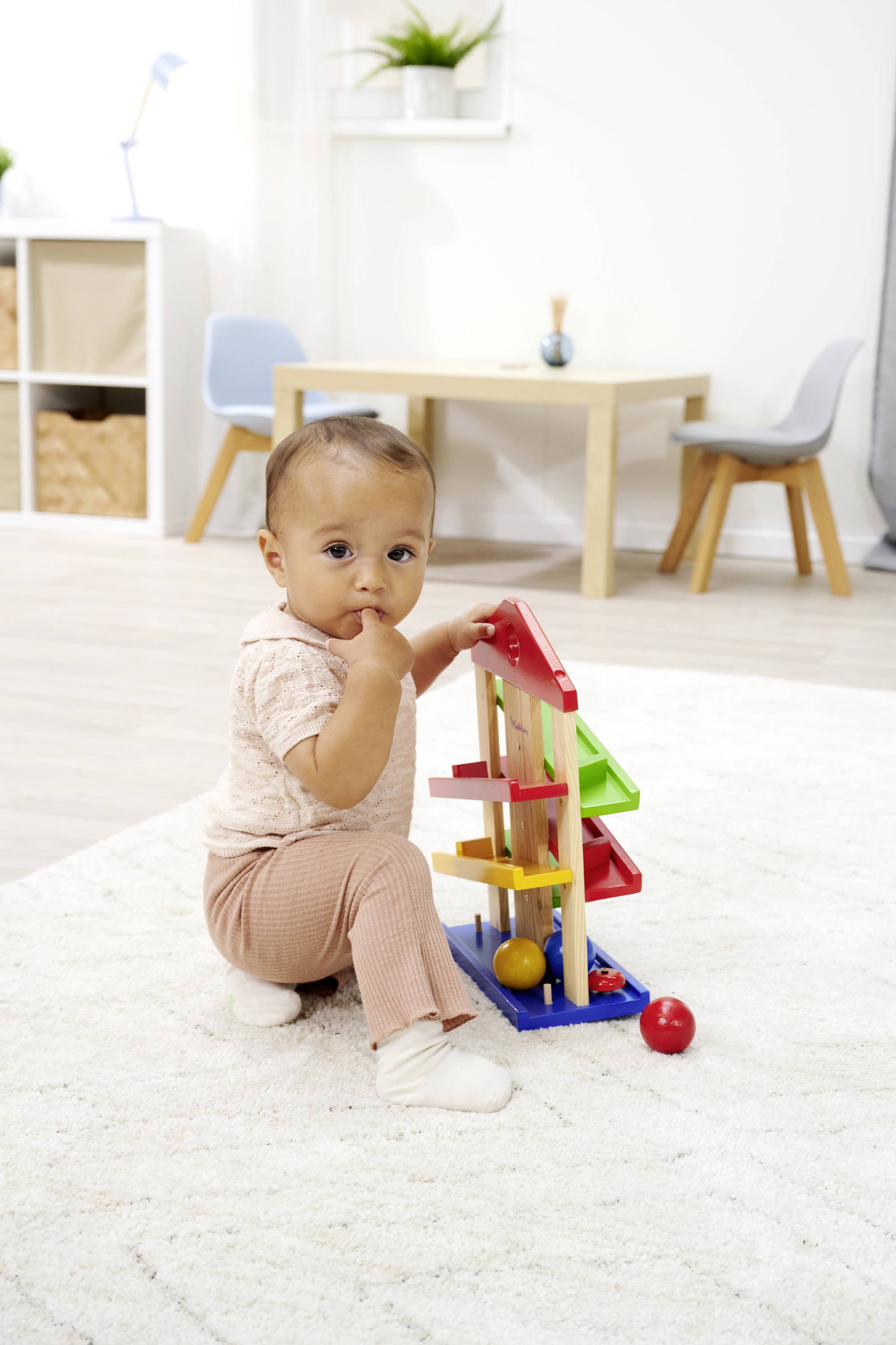 A toddler sits on a carpet and plays with a colourful marble run toy in a brightly decorated room.